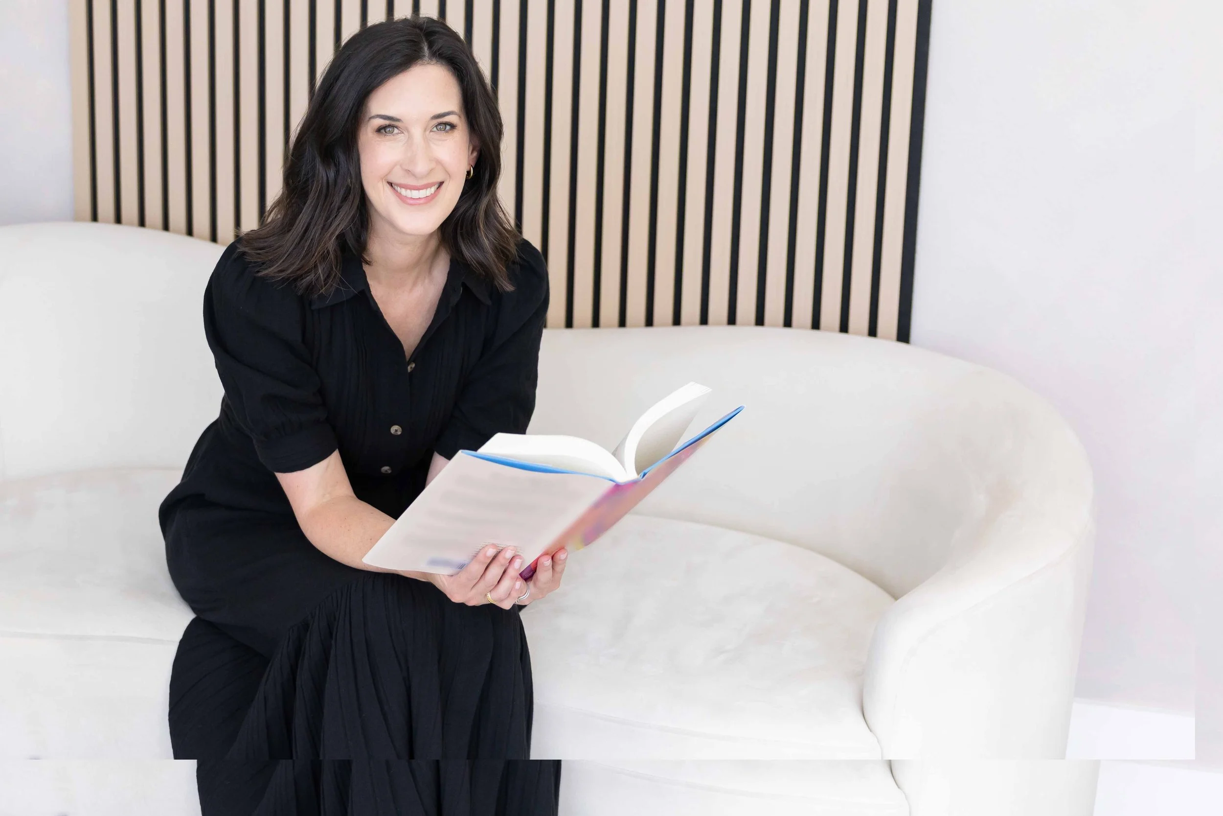 A woman with shoulder-length dark hair, wearing a black shirt and black skirt, sitting on a white curved bench and reading a book with a colorful cover. The background features a wall with black vertical slats.
