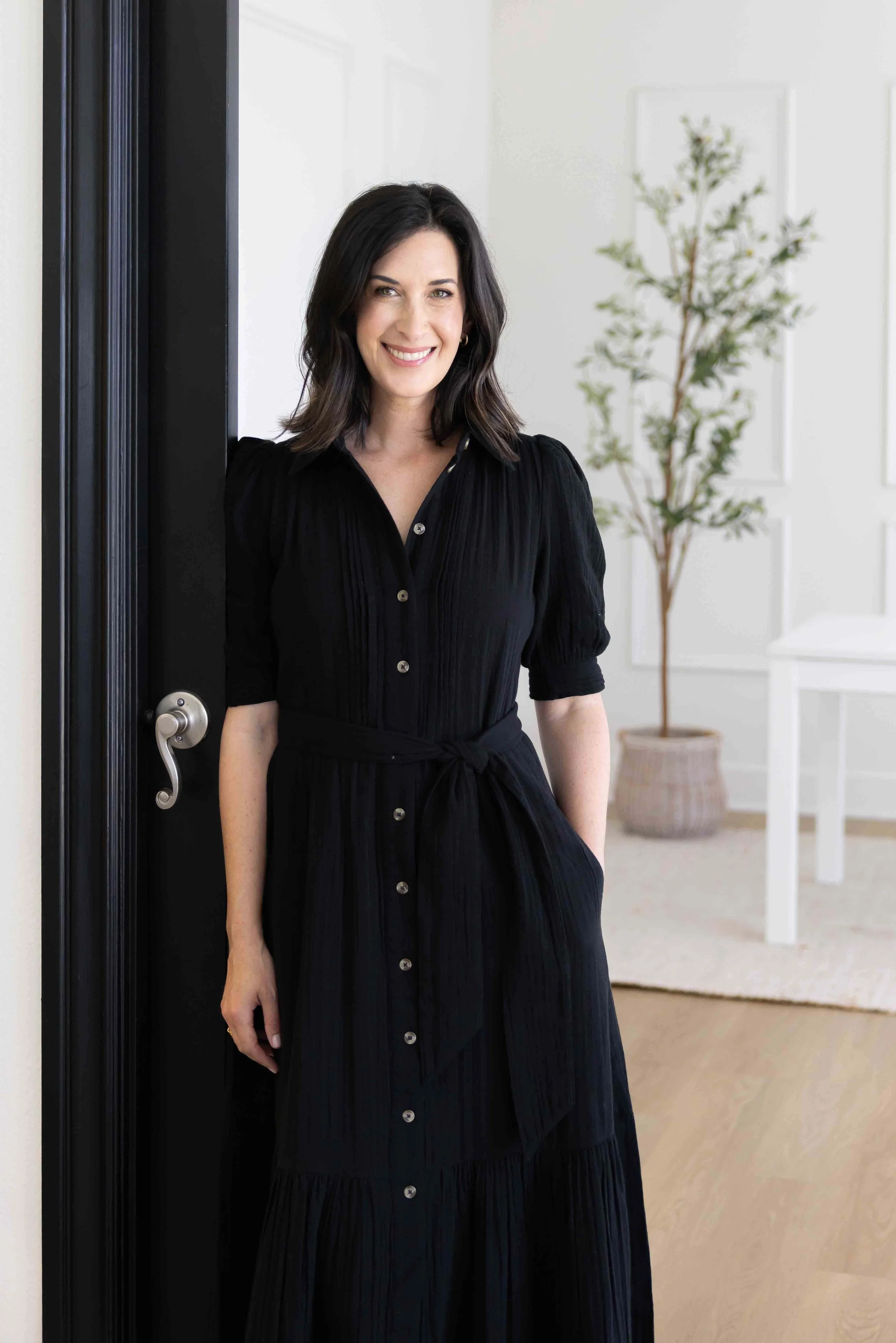 A woman with shoulder-length dark hair, smiling, wearing a black dress with buttons, standing in a doorway with a modern interior background featuring a potted plant and white wall decor.