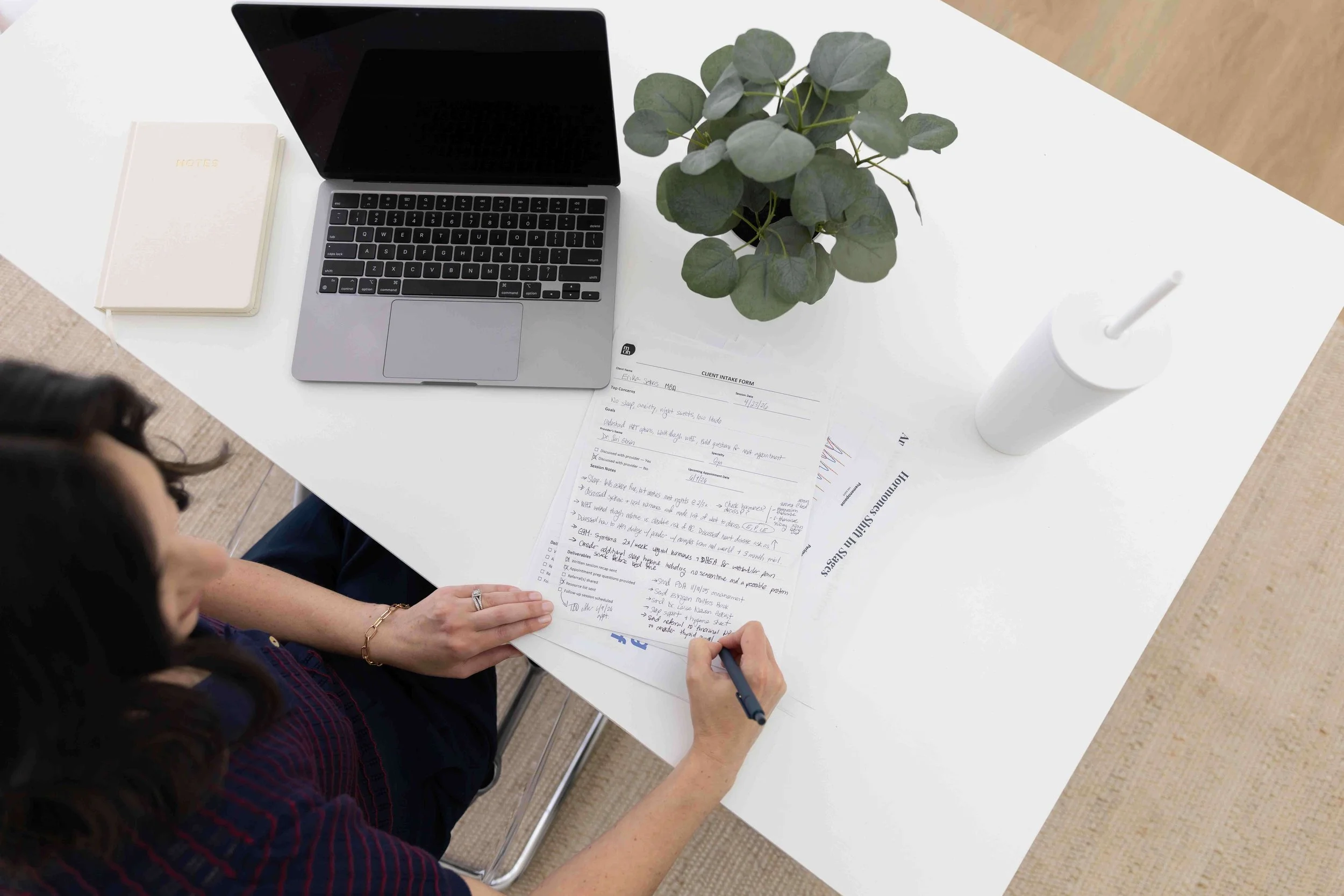 Overhead view of a person sitting at a white table, writing on a handwritten form, with a laptop, a plant, a closed notebook, and a white cup with a straw on the table.