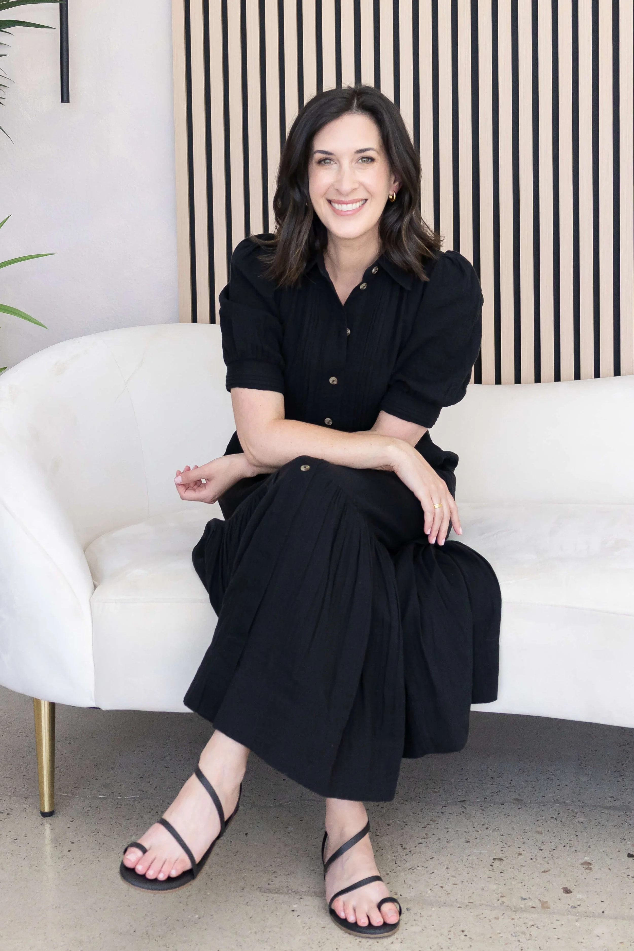 Woman with shoulder-length dark hair, wearing a black dress and black strappy high heels, sitting on a white sofa in a modern interior with a wooden slat wall background.