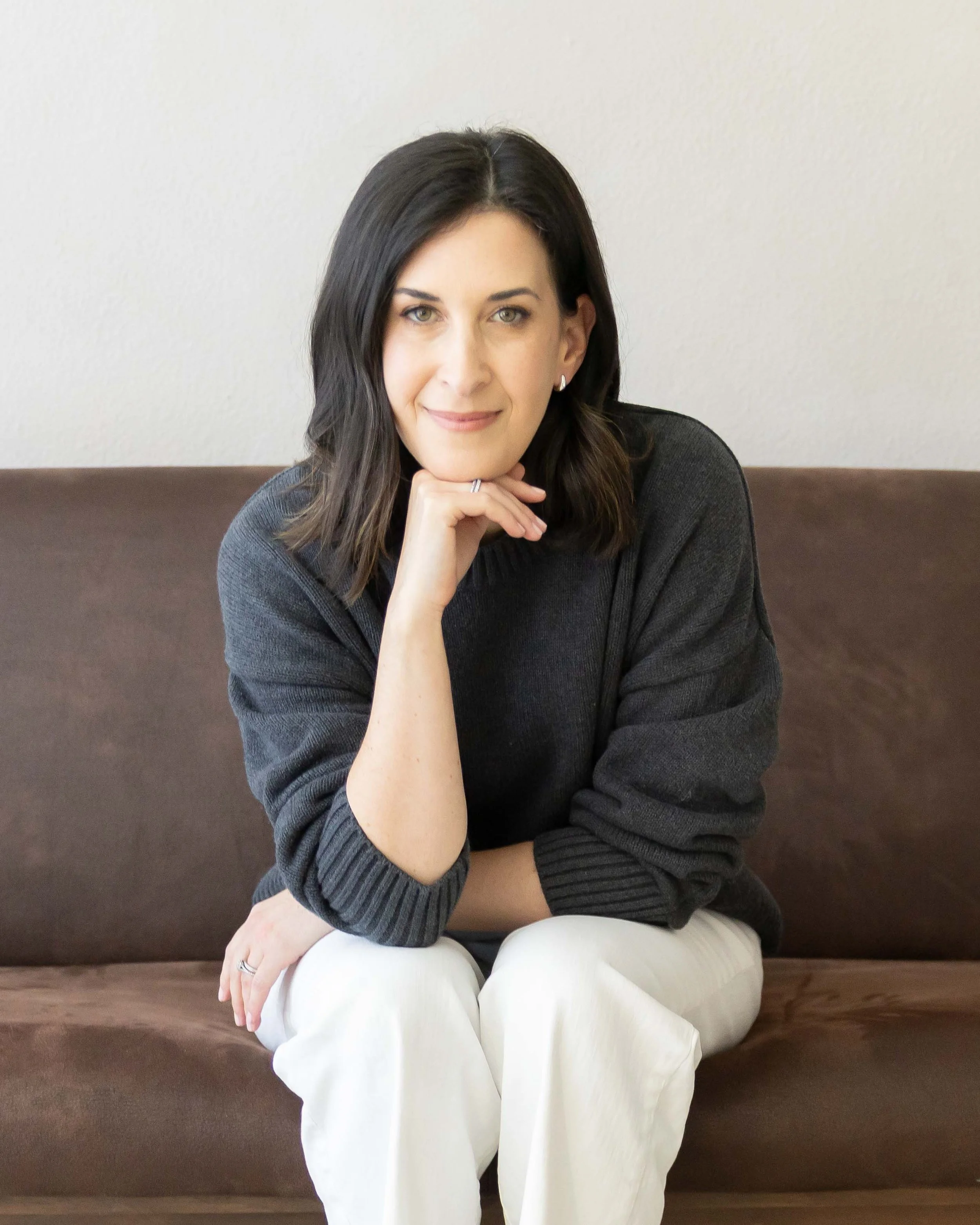 A woman with dark brown hair, light-colored eyes, and a subtle smile, seated on a brown bench against a plain white wall, resting her chin on her hand, wearing a dark sweater and light-colored pants.