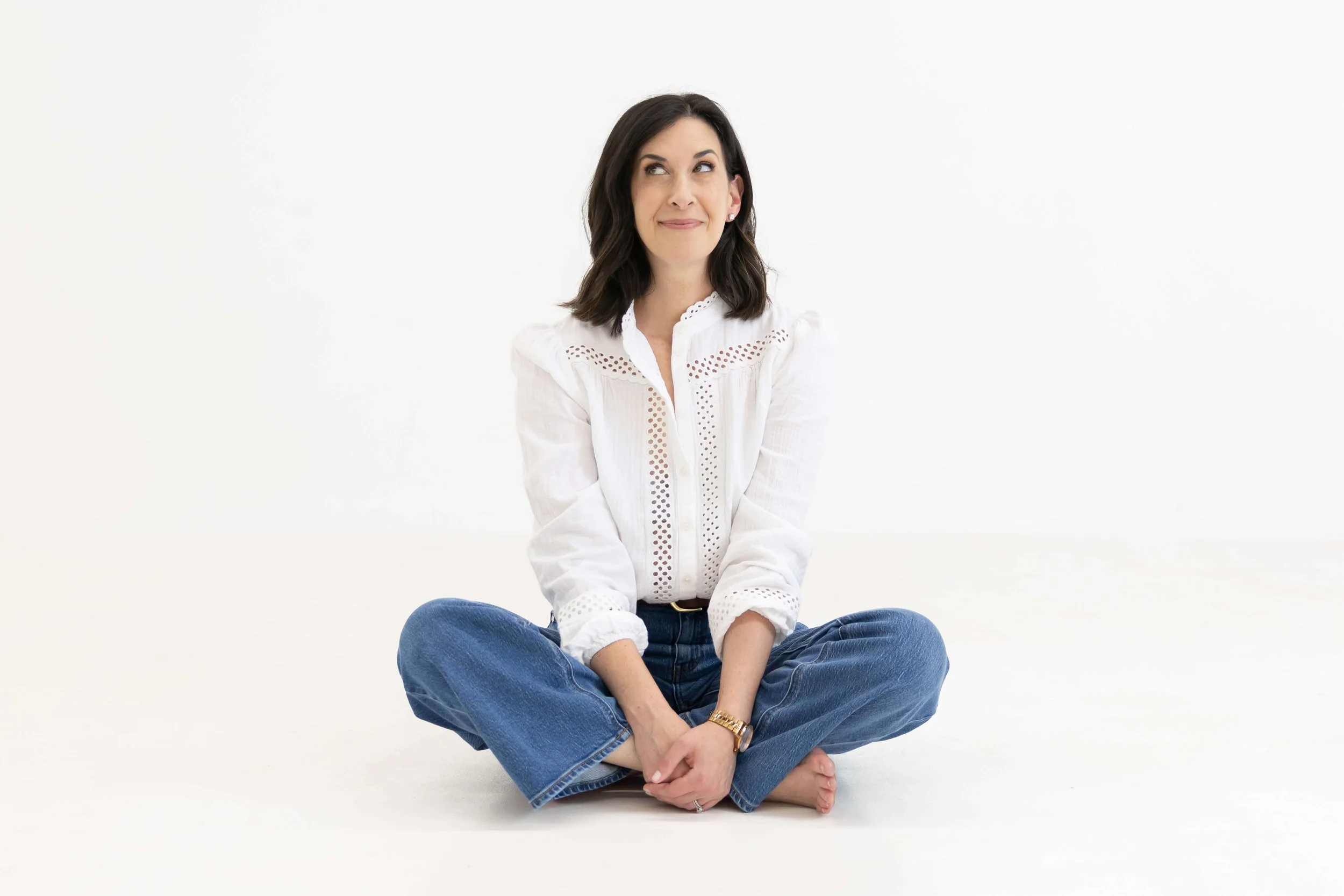 A woman with dark hair, wearing a white blouse and blue jeans, sitting cross-legged on the floor with a neutral background.