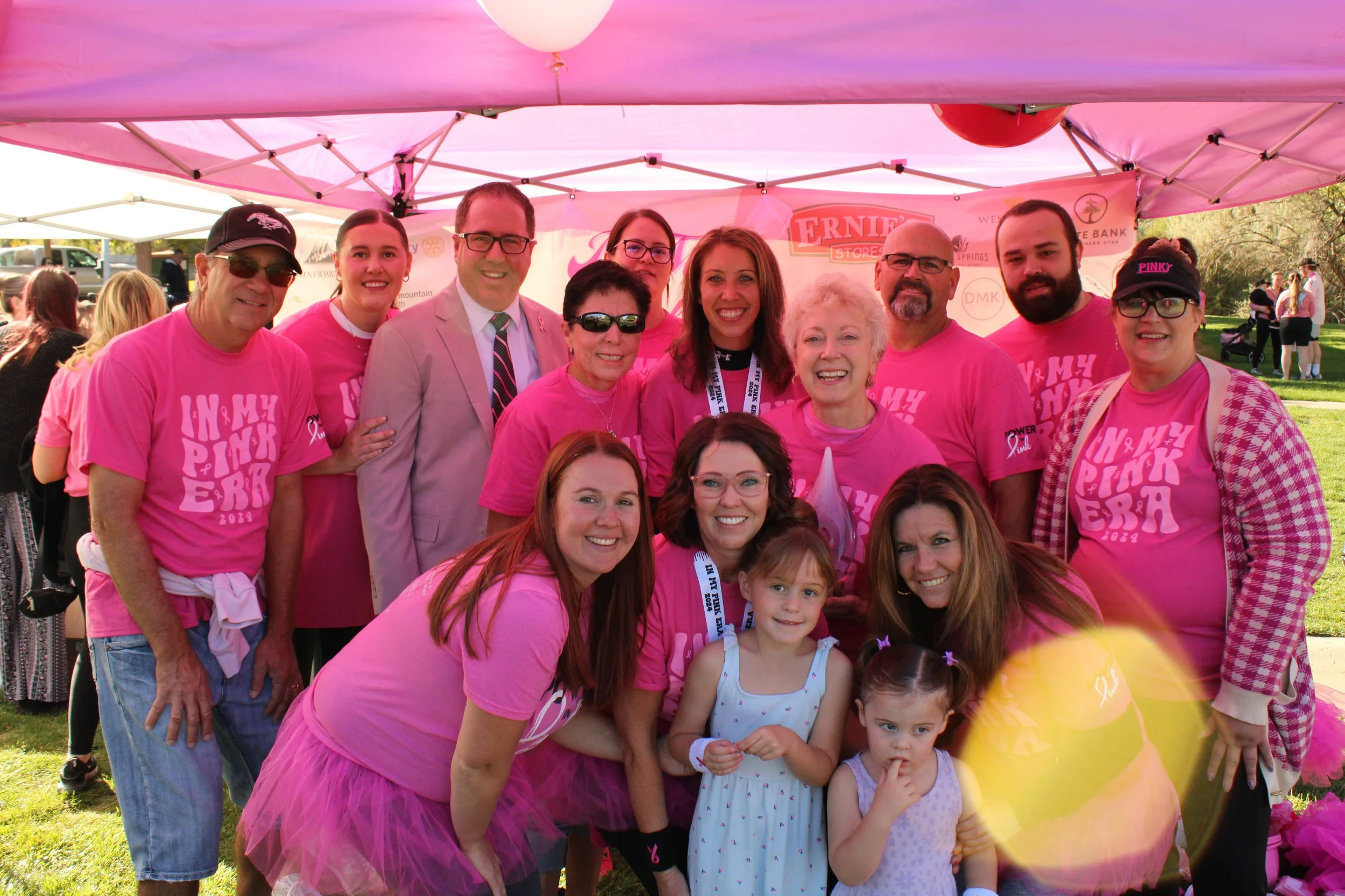 Group of people wearing pink shirts participating in a charity event, standing under a pink tent outdoors, with balloons and other people in the background.