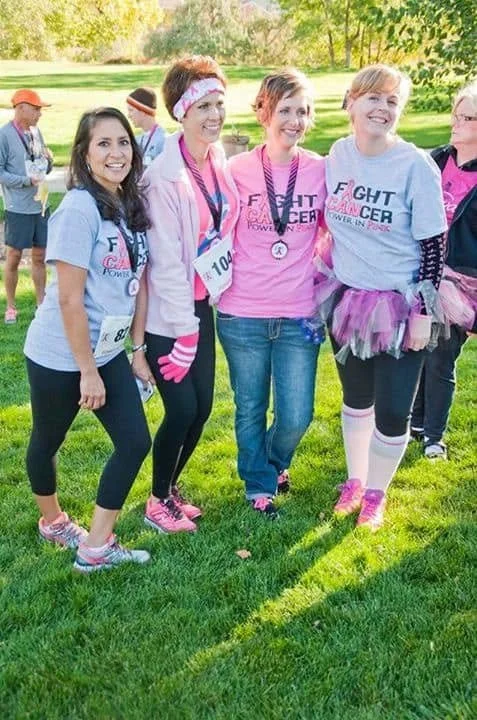 Group of women at a breast cancer awareness event, wearing race shirts and medals, standing together outdoors.