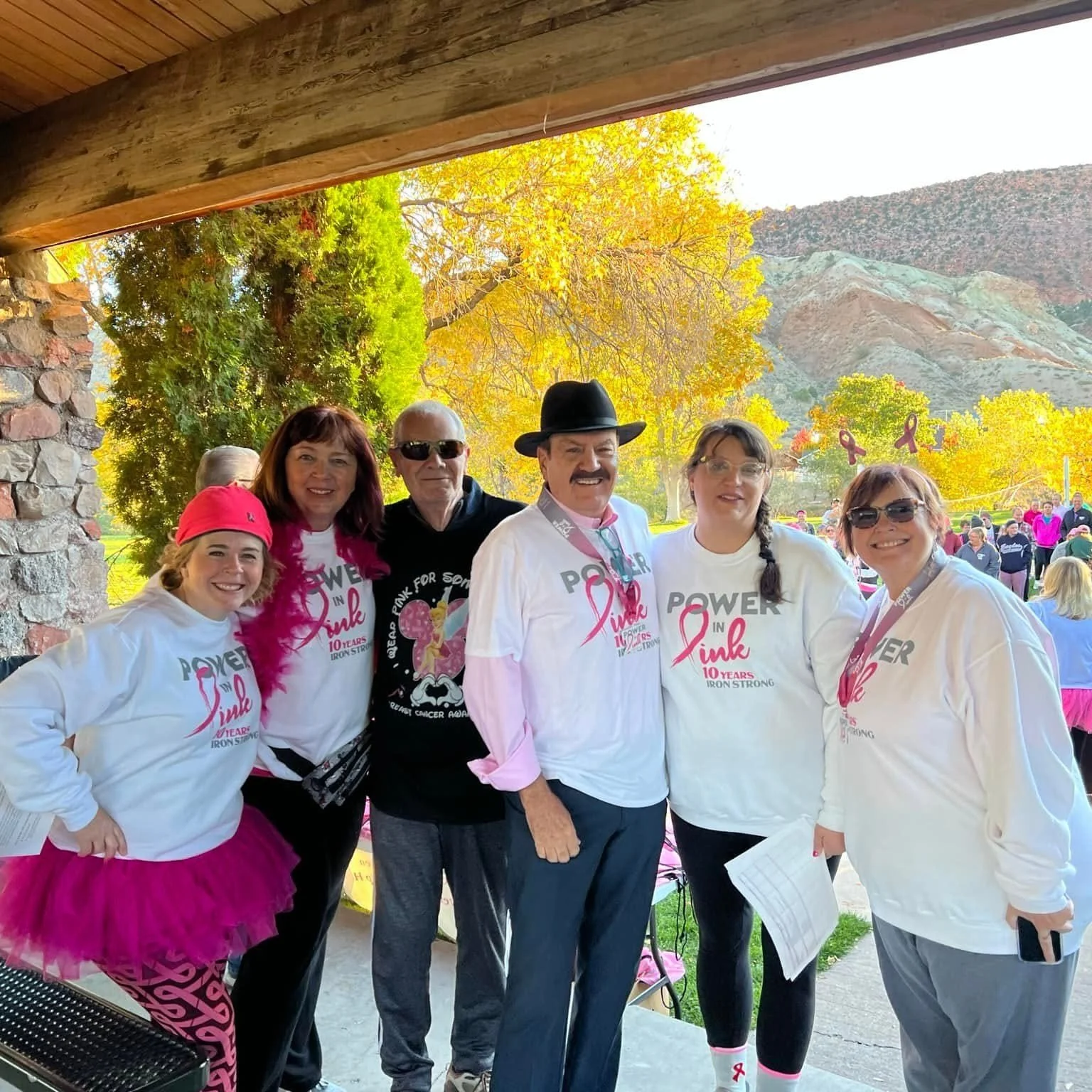 Group of six people standing together outdoors, wearing white shirts with pink ribbons that say 'Power in Pink 10 Years Iron Strong,' celebrating a breast cancer awareness event, with autumn trees and mountains in the background.
