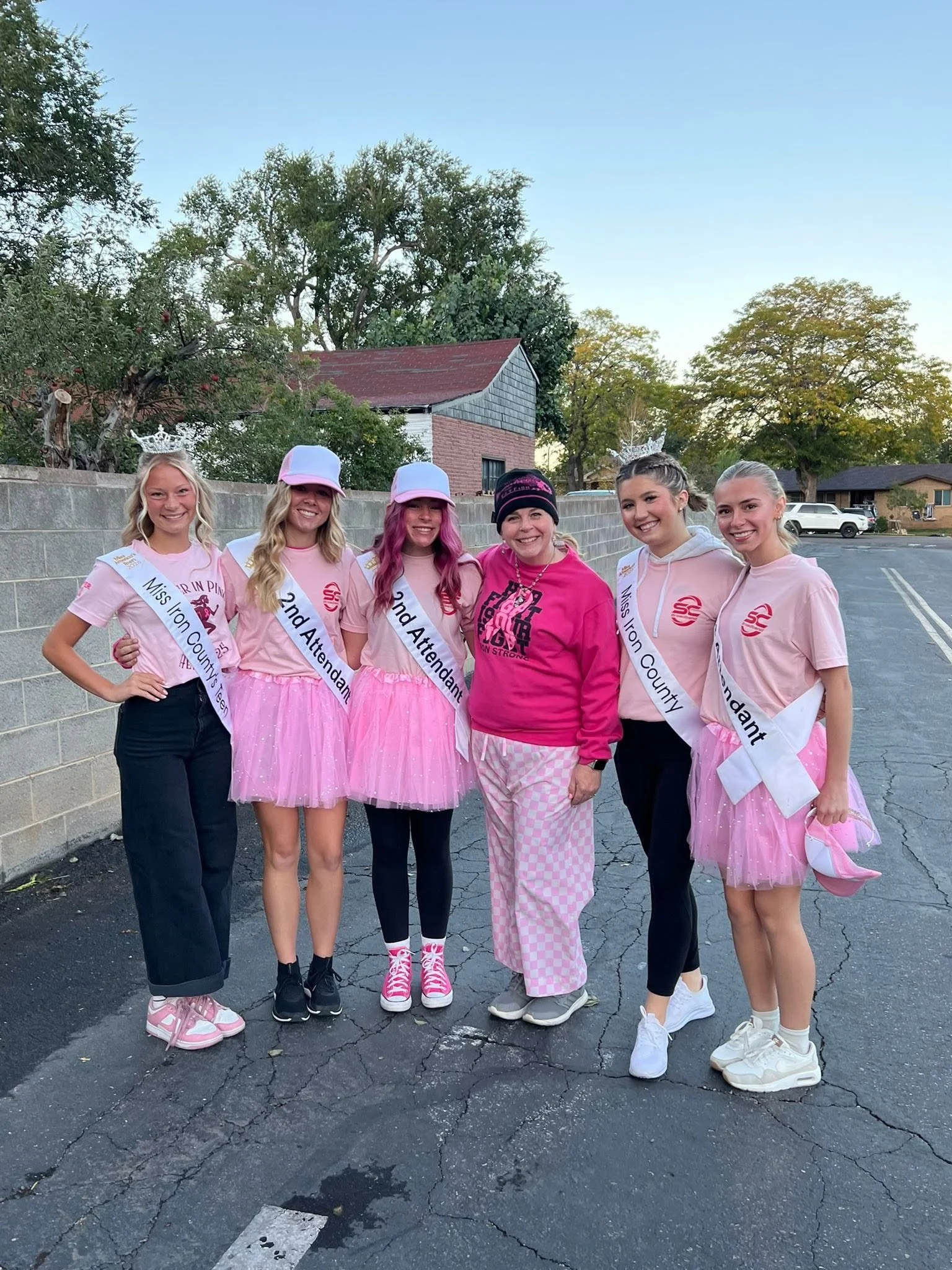 Group of six women with crowns and sashes, standings outdoors in front of a brick wall, wearing pink dresses and casual clothing, smiling for a picture.