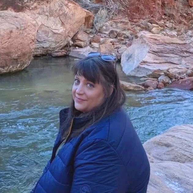 A woman with dark hair and bangs, wearing a blue jacket and sunglasses on her head, sitting by a rocky stream in a canyon.