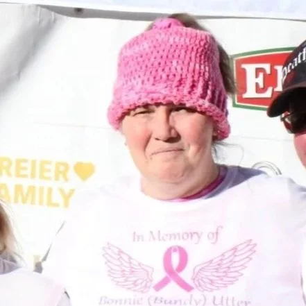 Woman wearing a pink knitted hat and a white shirt with a pink breast cancer awareness logo, standing outdoors.
