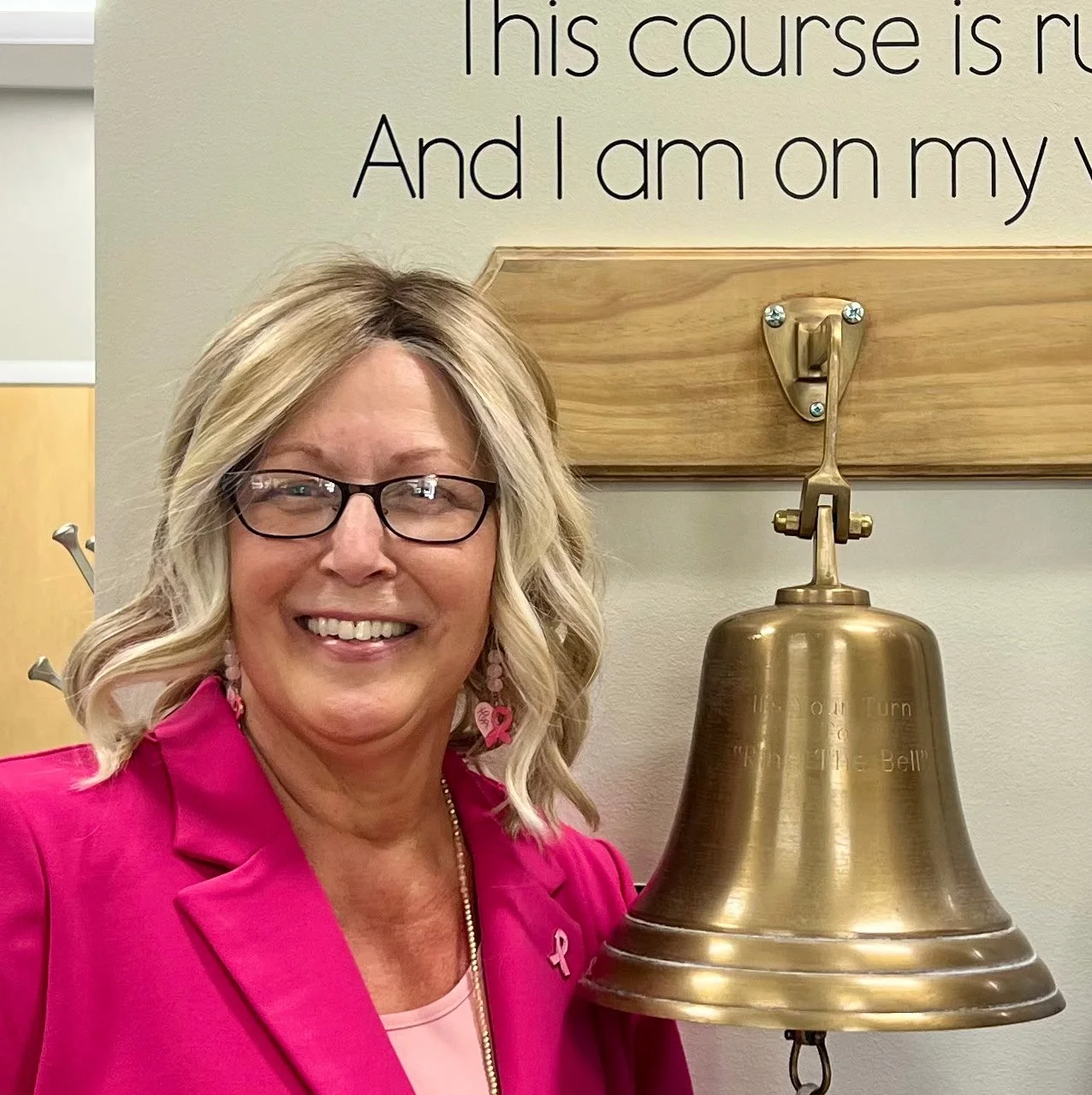 A smiling woman with blonde hair, glasses, and pink earrings, wearing a pink blazer, standing next to a large brass bell on a wall hook, in front of a beige wall with black text.