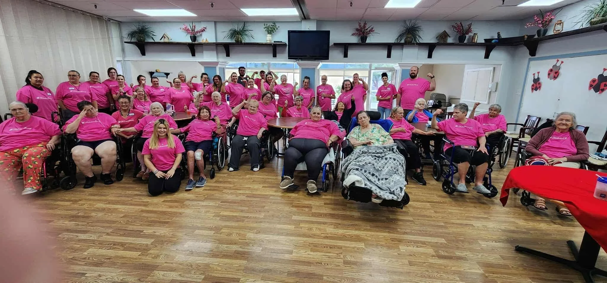 A large group of people, most wearing pink shirts, gathered in a room with wooden flooring and decorated with plants and wall art. Some people are seated in wheelchairs, others standing, posing for a group photo.