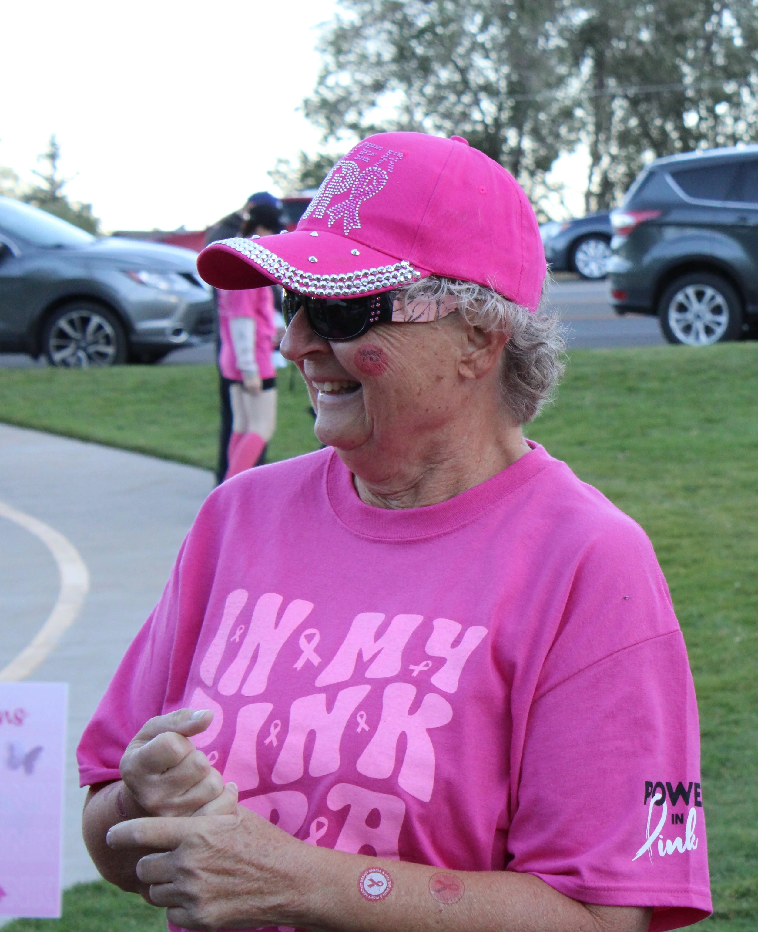 Profile of a smiling elderly woman wearing a pink cap with rhinestones, sunglasses, and a pink shirt at a breast cancer awareness event in an outdoor park.