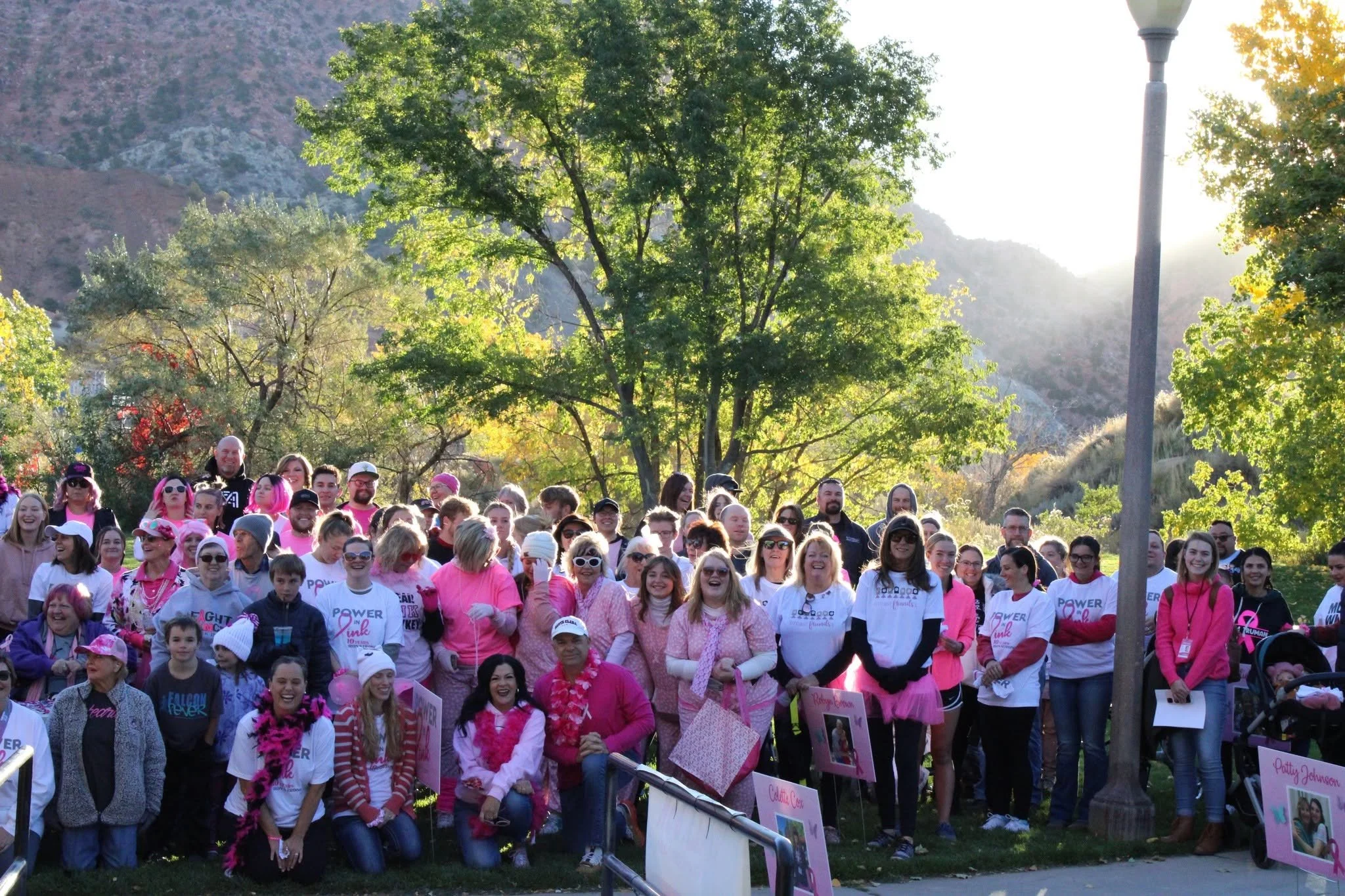 Large group of people gathered outdoors for a walk or event, many wearing pink and white, with some holding signs or pictures, surrounded by trees and natural scenery.