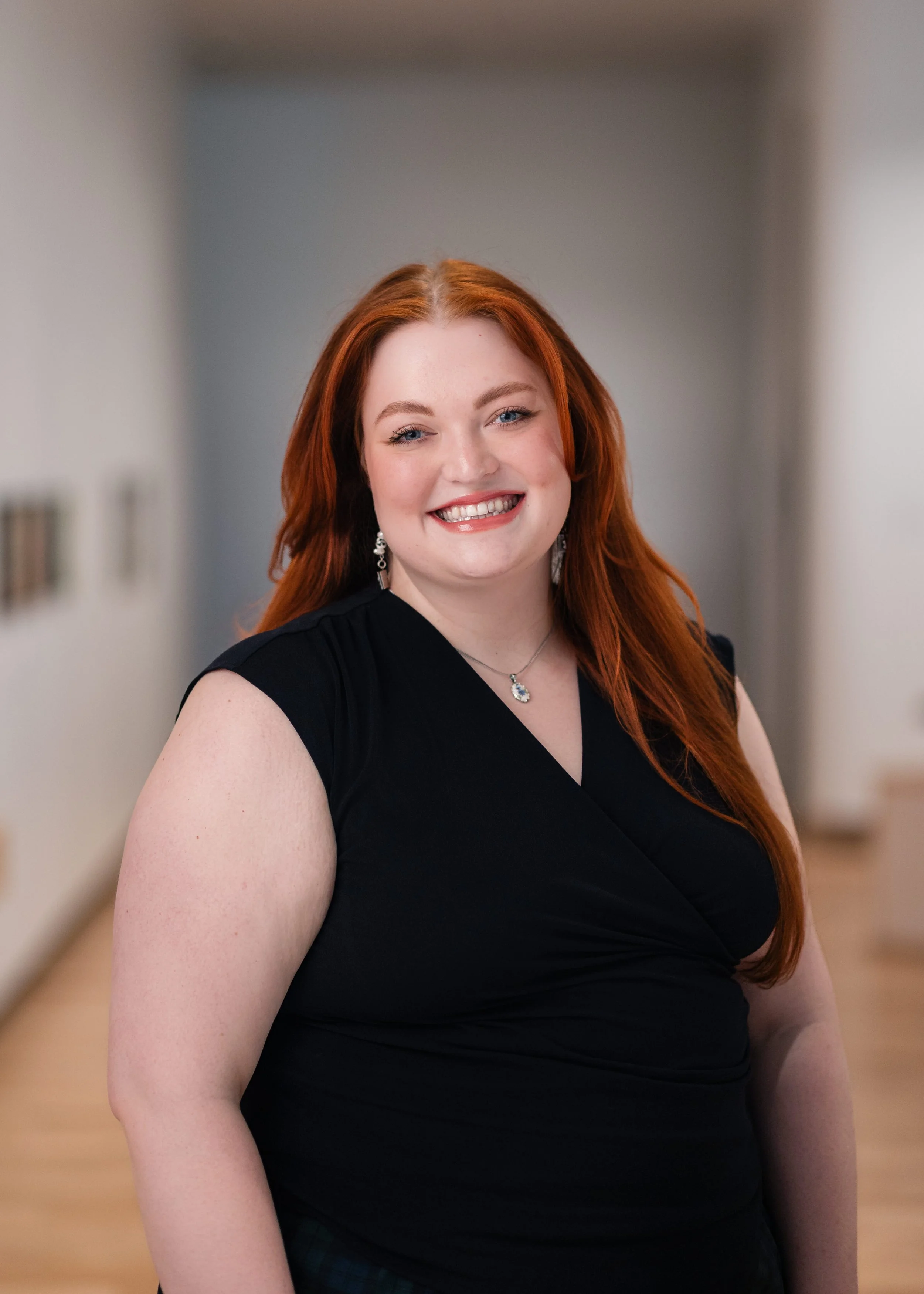A woman with red hair and fair skin, smiling, wearing a black dress and jewelry, standing indoors with a blurred background.