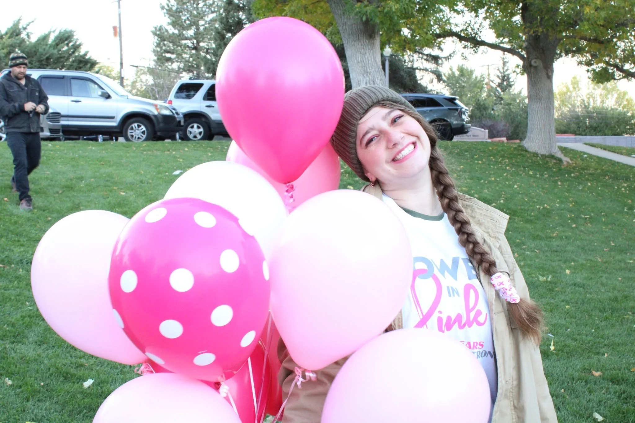 Young woman smiling outdoors holding pink balloons, wearing a beanie and beige jacket, in a park with trees and cars in the background.