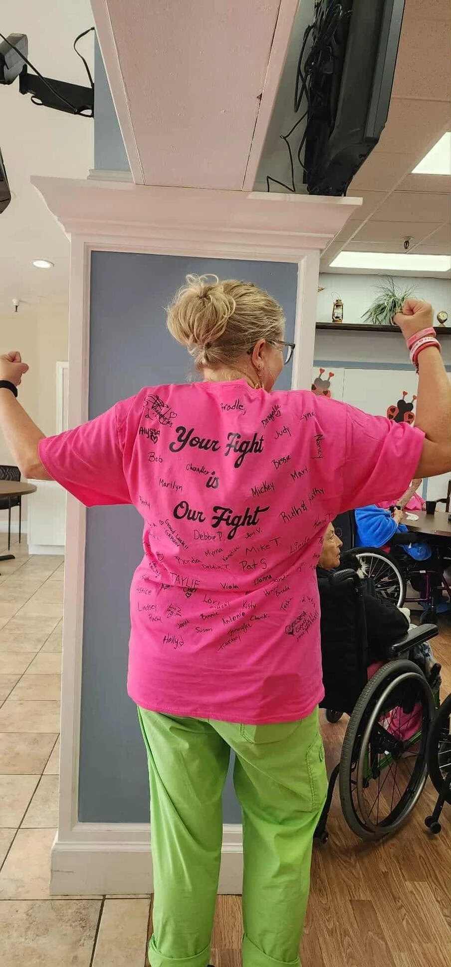 A woman in a pink shirt with signatures and messages stands with her arms raised, flexing her muscles. She is in a room with a blue wall behind her and other people, including a woman in a wheelchair, in the background.