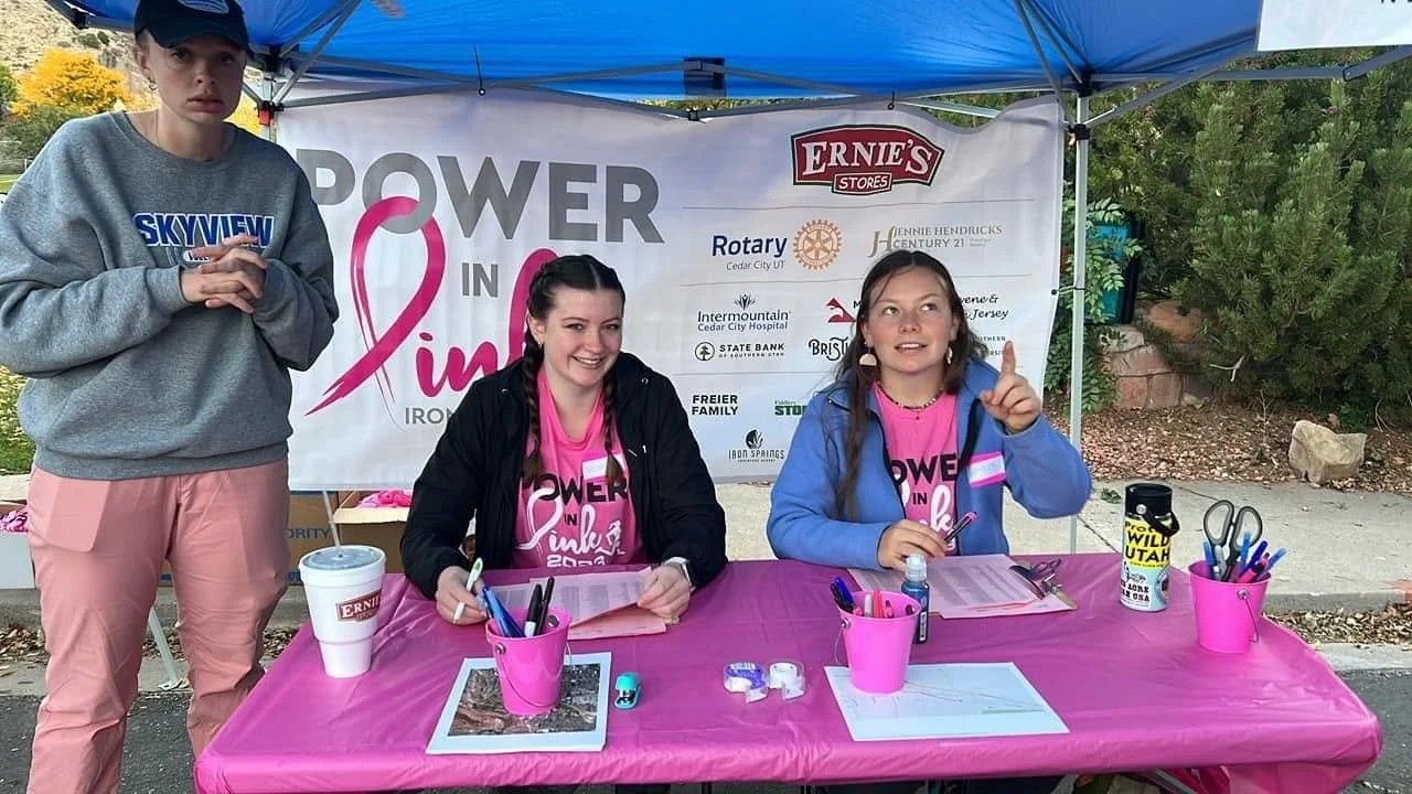 Three young women at a table under a blue canopy, participating in a fundraising event called 'Power in Pink' for breast cancer awareness. Two are sitting at the table, smiling, while one is standing. The table has pink tablecloths and various office supplies.