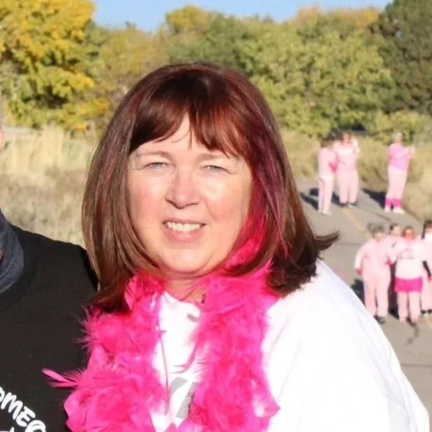 Woman with red hair and pink feather boa smiling outdoors with trees and people in pink clothing in the background.