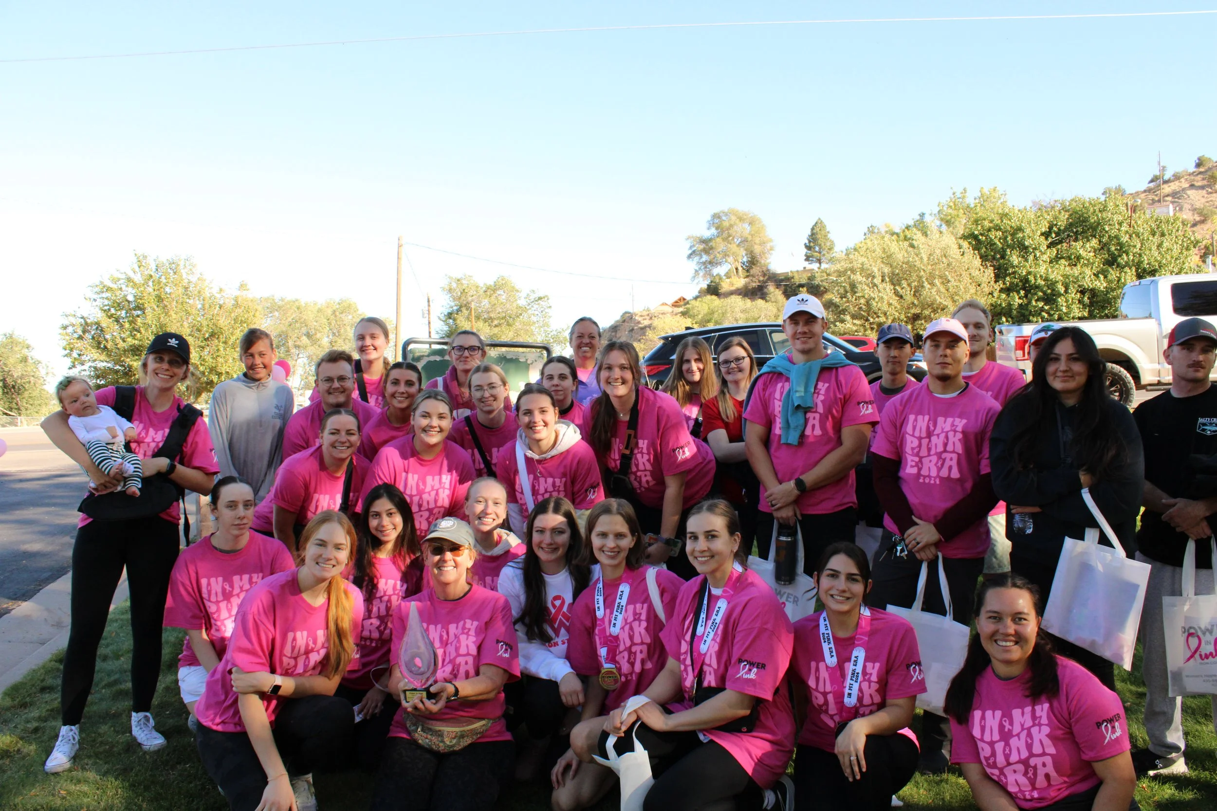 A large group of people, mostly women, wearing pink shirts, gathered outdoors for a charity event, with some holding medals and trophies, and wearing pink ribbons for breast cancer awareness.