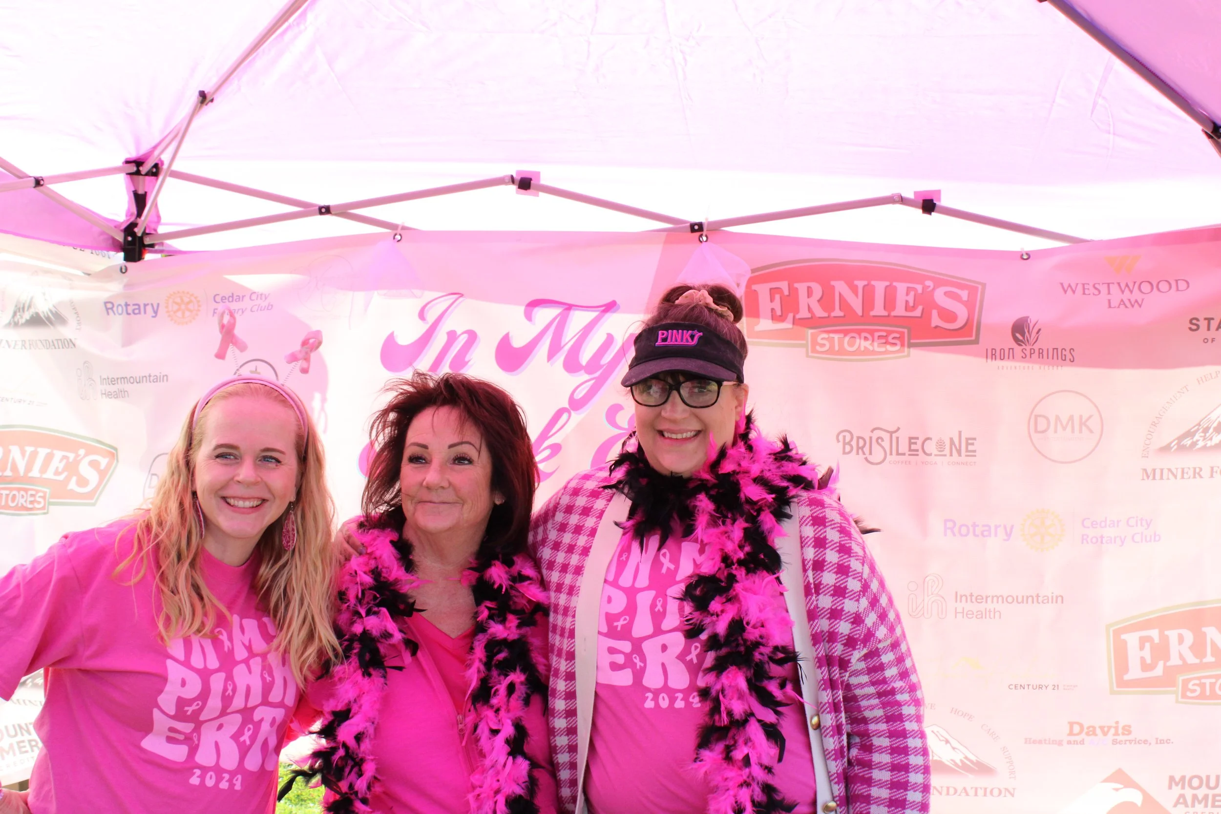 Three women standing together under a pink event tent, wearing pink shirts and accessories supporting breast cancer awareness at a charity event.