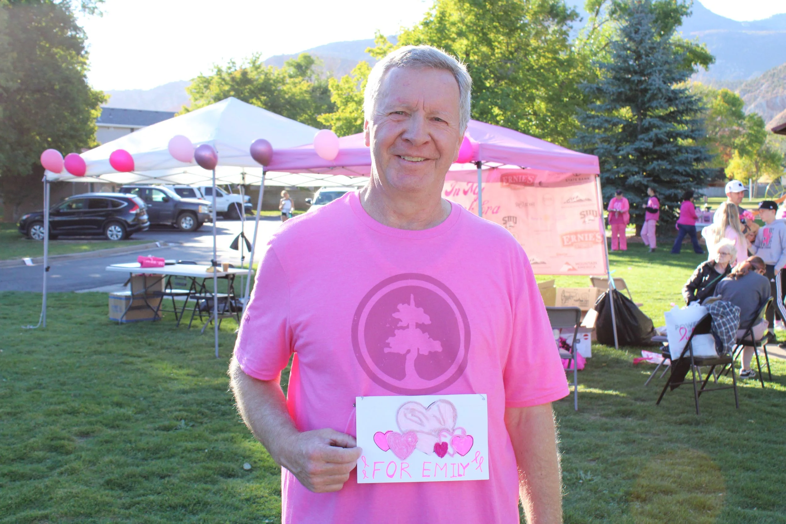 A smiling man standing outdoors at a fundraising event, wearing a pink t-shirt and holding a sign with pink hearts and the words "For Emily." In the background, there are tents decorated with pink and purple balloons, other people participating in the event, and a scenic area with trees and mountains.