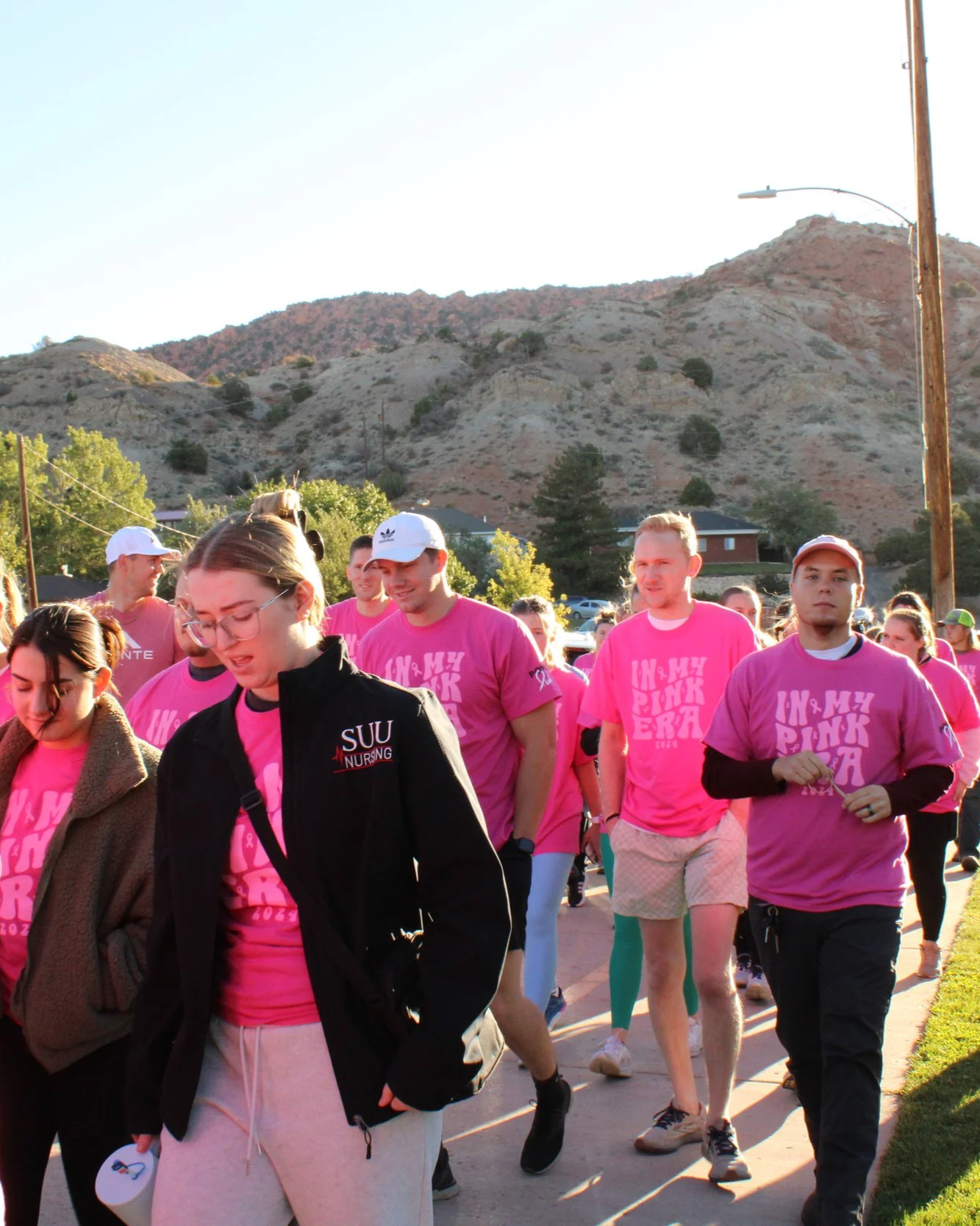 A group of people participating in a walkathon outdoors, many wearing pink shirts that say 'In My Pink Era.' They are walking along a sidewalk with a mountain in the background and a clear sky.