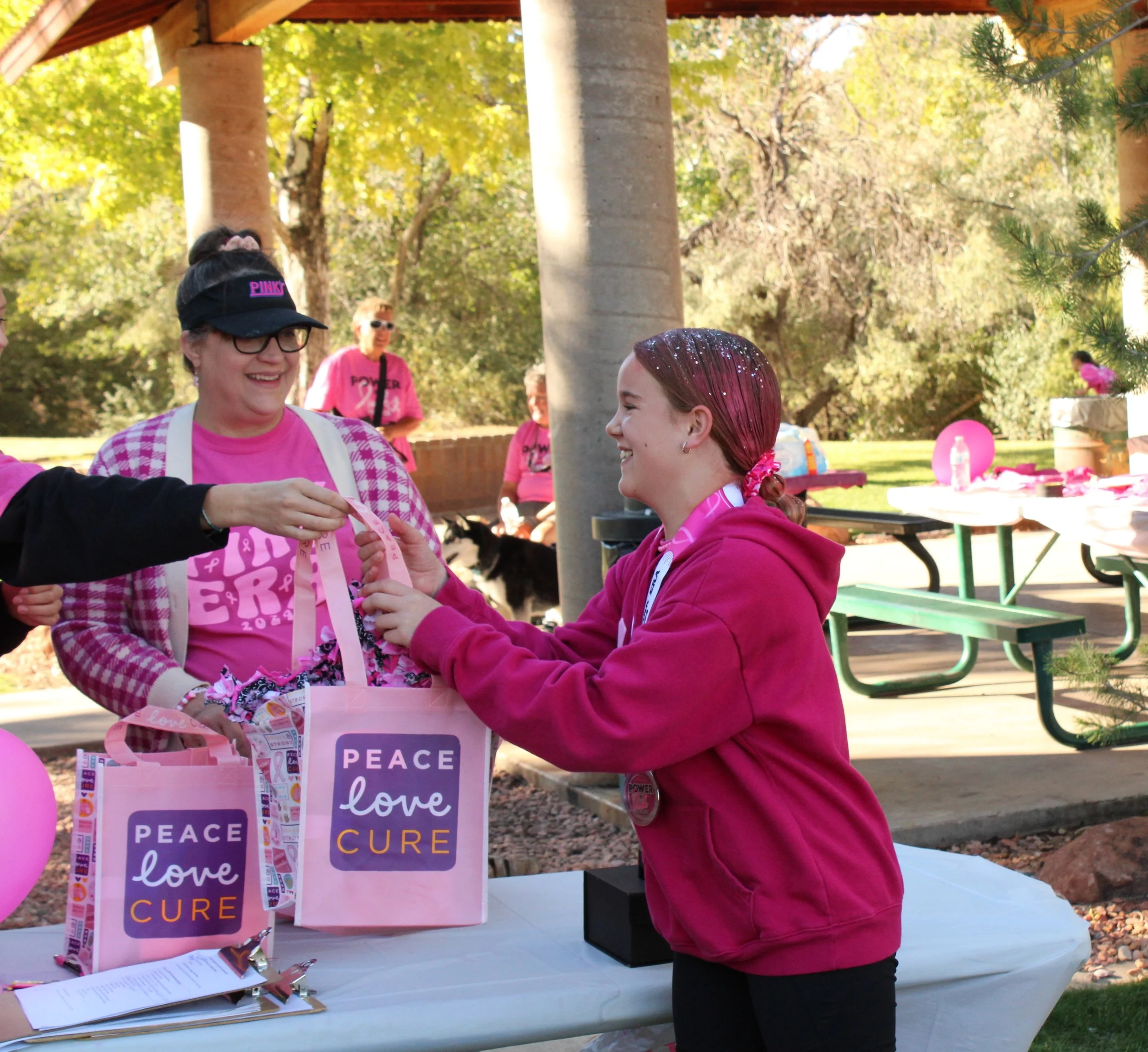Two women at a table during an outdoor breast cancer awareness event. The woman on the left is wearing a pink hat, glasses, and a pink checkered shirt, smiling as she receives a ribbon. The woman on the right has red hair tied back with a pink scrunchie, wearing a pink hoodie, and is handing the ribbon to the other woman. The table has pink balloons and pink tote bags with messages like "peace", "love", and "cure." In the background, there are trees with green and yellow leaves and other people wearing pink.