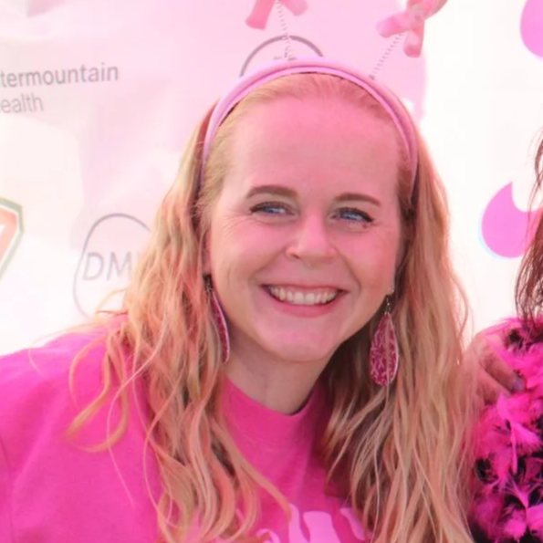 A smiling woman with long blonde hair, wearing pink headband and pink earrings, dressed in a pink shirt, at a pink-themed event or celebration.
