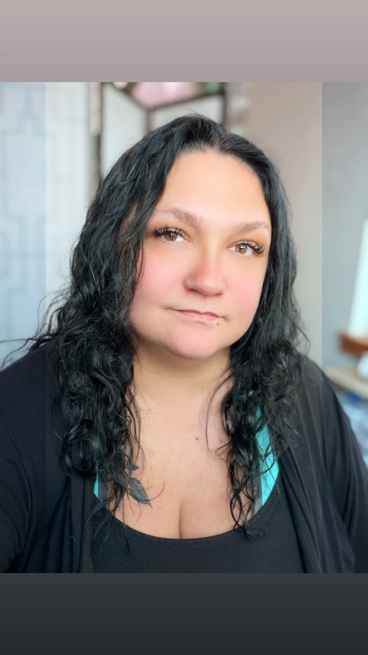 Close-up of a woman with long curly black hair, hazel eyes, and nose piercings, wearing a black top with teal accents, indoors with a blurred background.