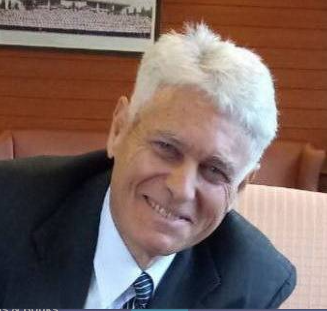 A smiling man with white hair wearing a dark suit and tie, sitting indoors with wood-paneled walls behind him.