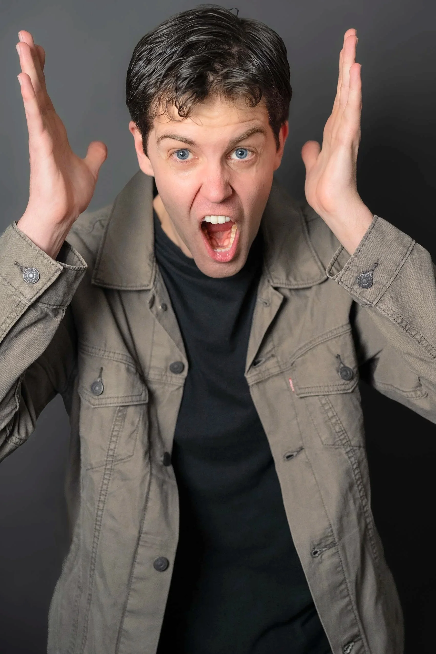 A man with short dark hair and blue eyes is making a yelling or shouting face, with his mouth open and eyes wide. He is holding both of his hands near his head, with palms facing outward. He is wearing a light gray jacket over a black shirt, against a plain dark background.