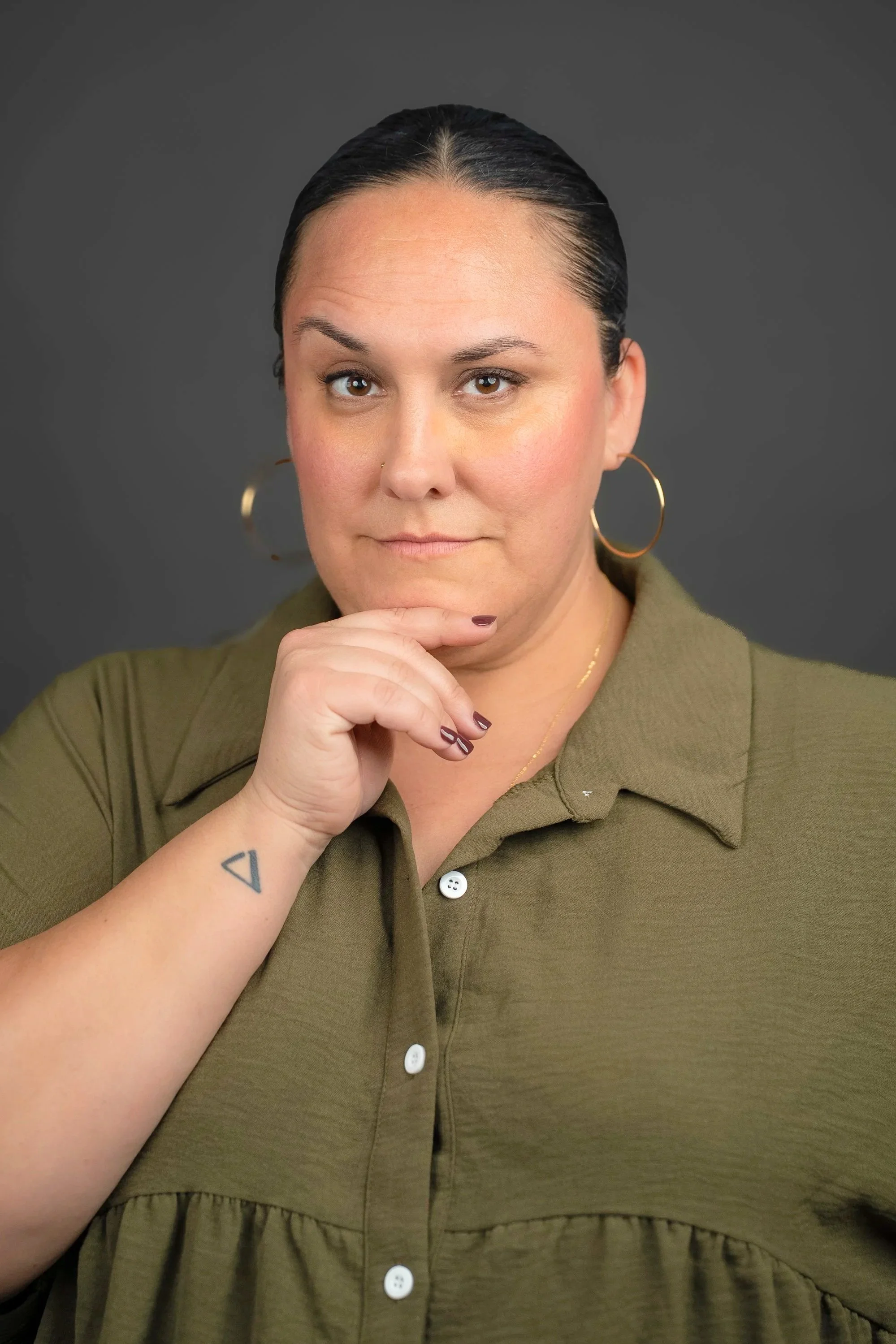 A woman with dark hair slicked back, wearing large hoop earrings and a green button-up top, rests her chin on her hand with a thoughtful expression against a gray background.