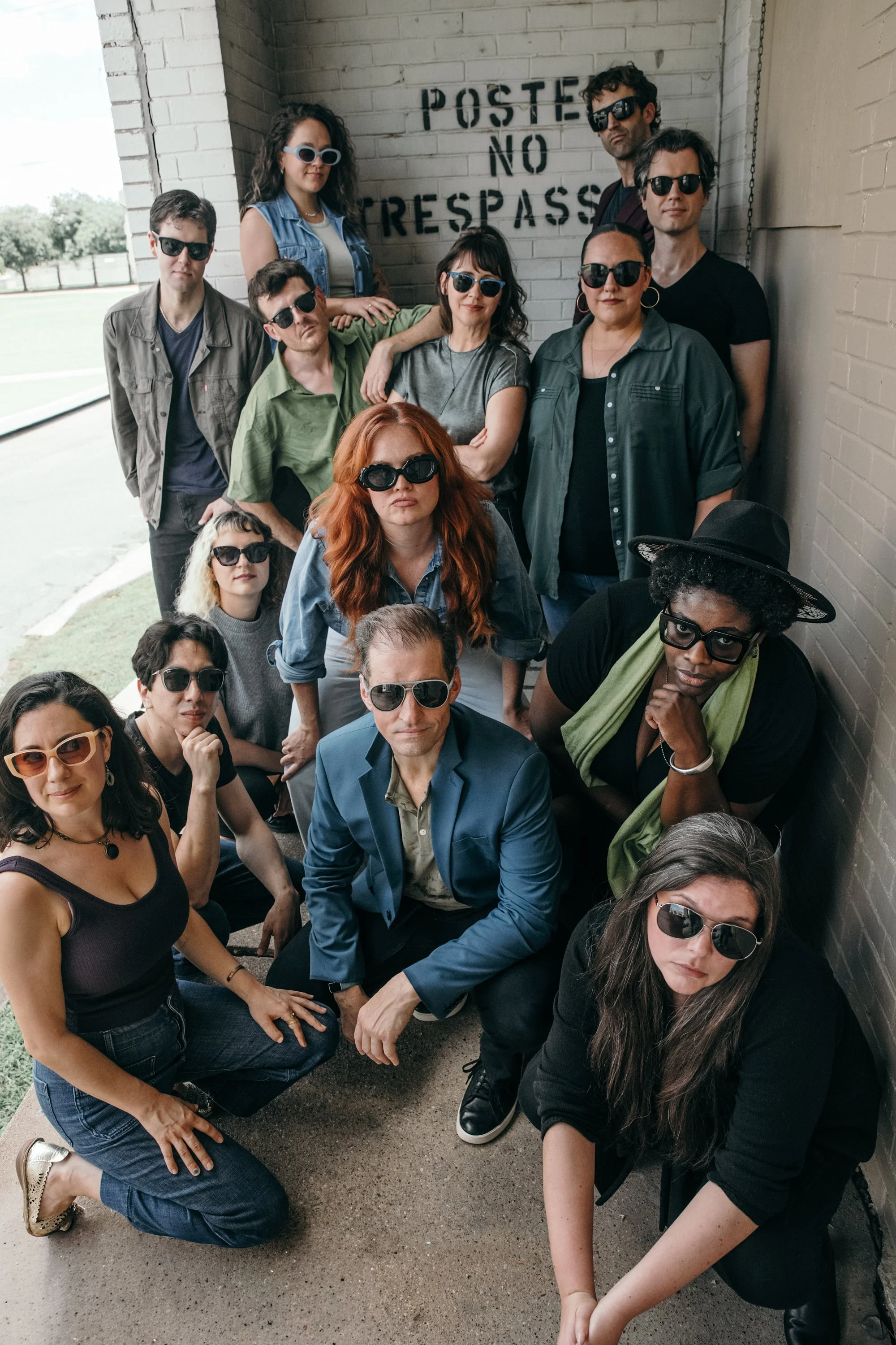 Group of diverse people wearing sunglasses, posing together outdoors against a wall.