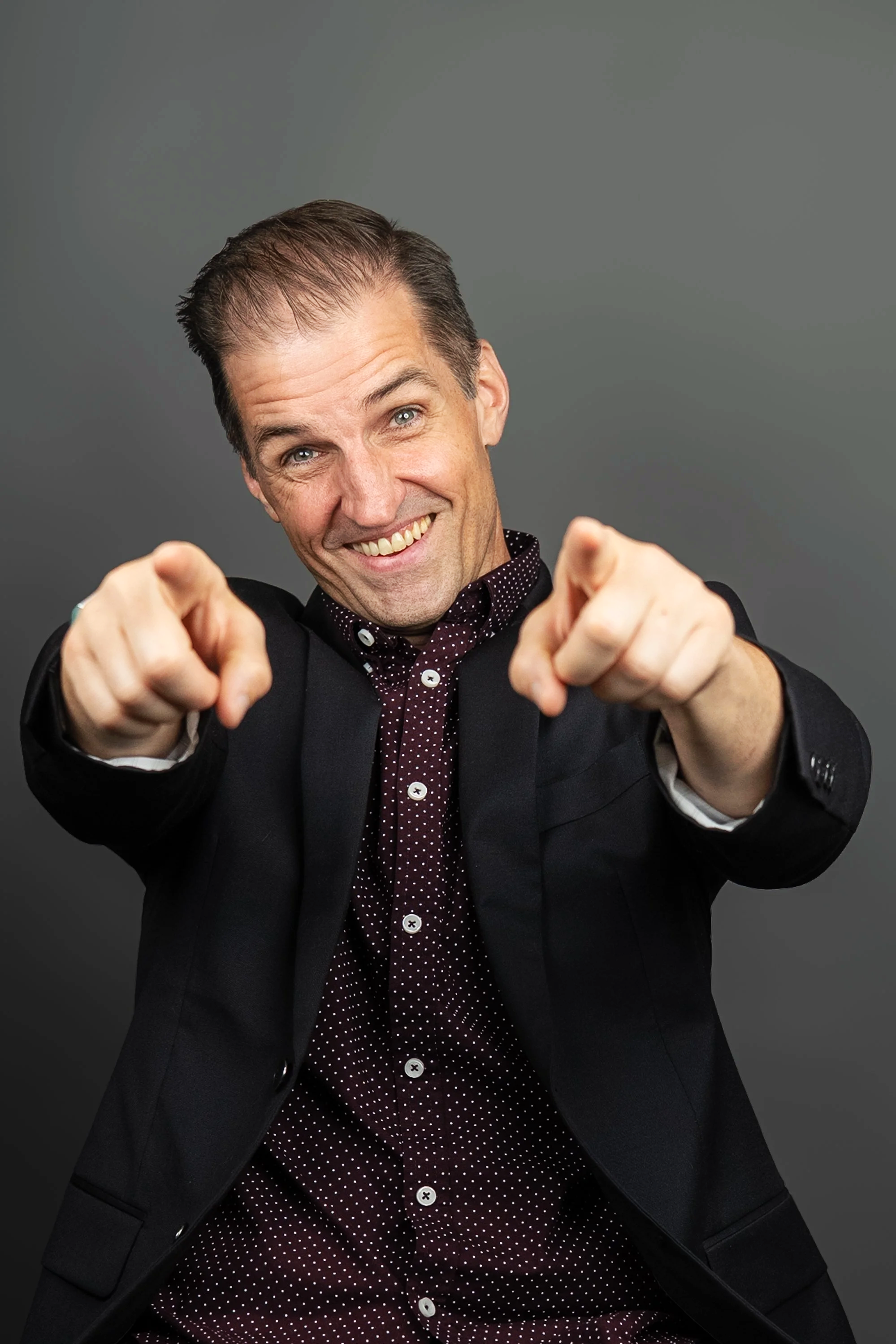 A cheerful man in a black suit and burgundy shirt with white polka dots is pointing at the camera with both hands, smiling, in front of a plain gray background pointing at the viewer.