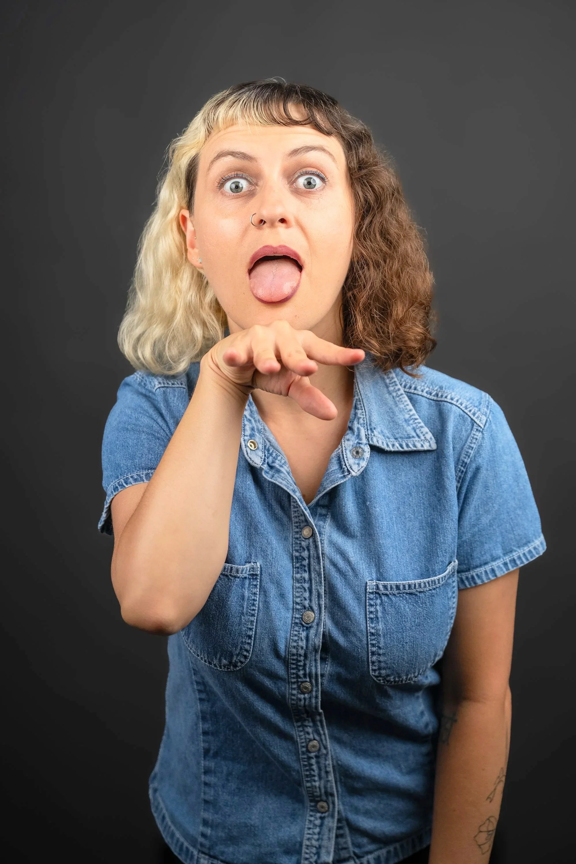 A woman with curly and straight half blonde and half brown hair, wearing a denim shirt, sticking out her tongue and holding her hand under her chin against a gray background.
