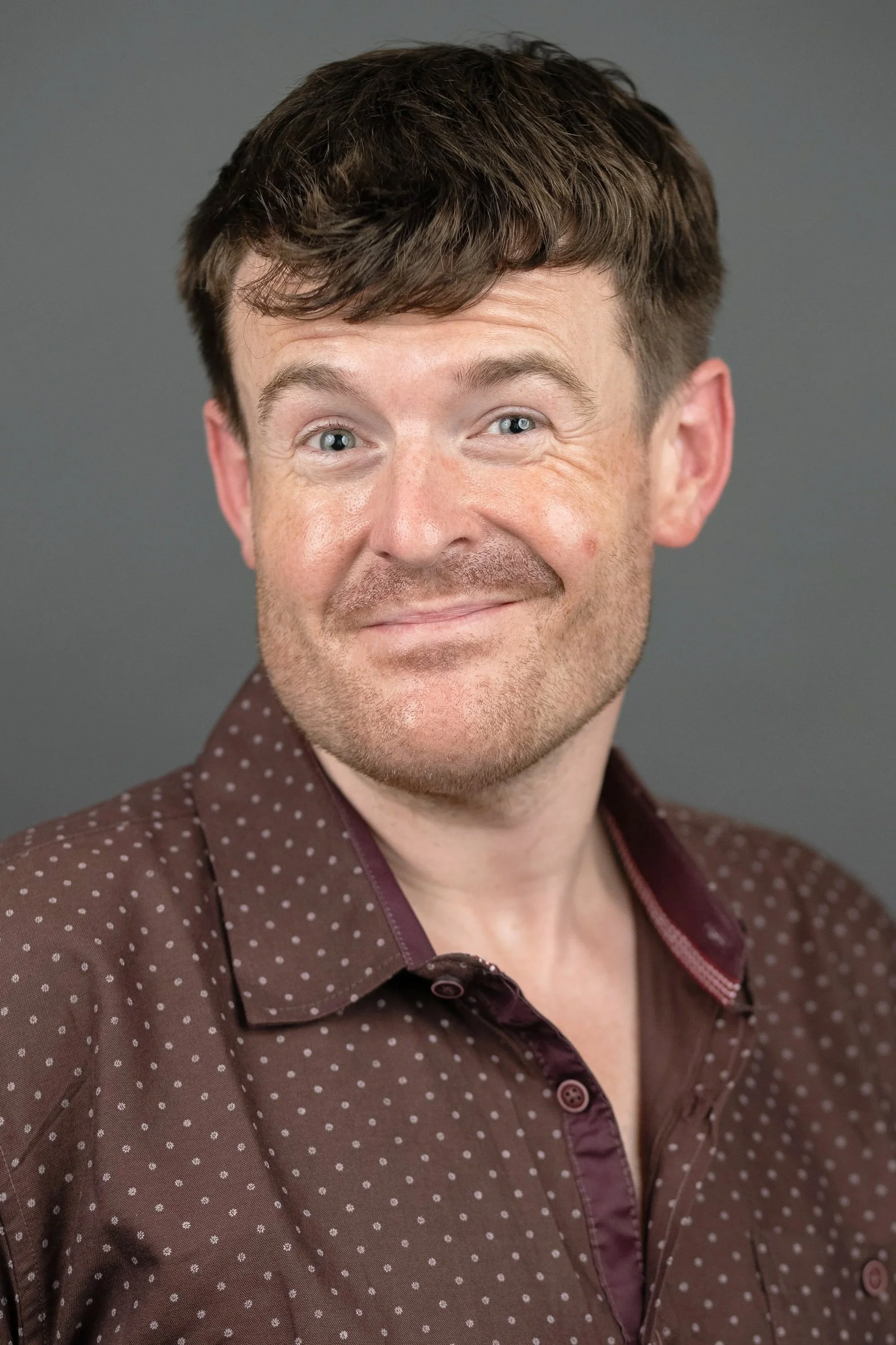A smiling man with short brown hair and light skin wearing a brown polka dot shirt against a plain gray background.