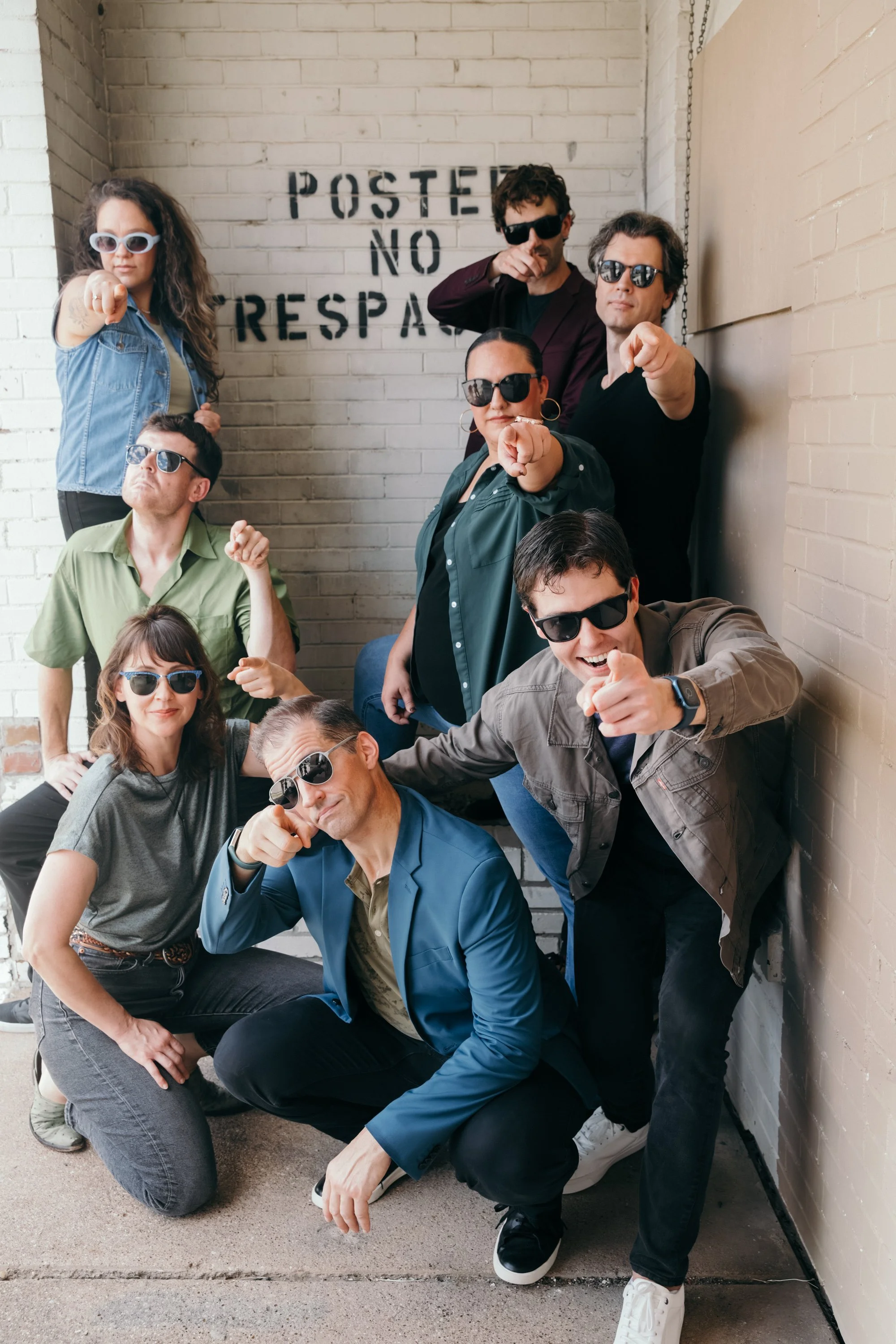Group of seven people wearing sunglasses, posing and pointing at the camera, against a white brick wall.