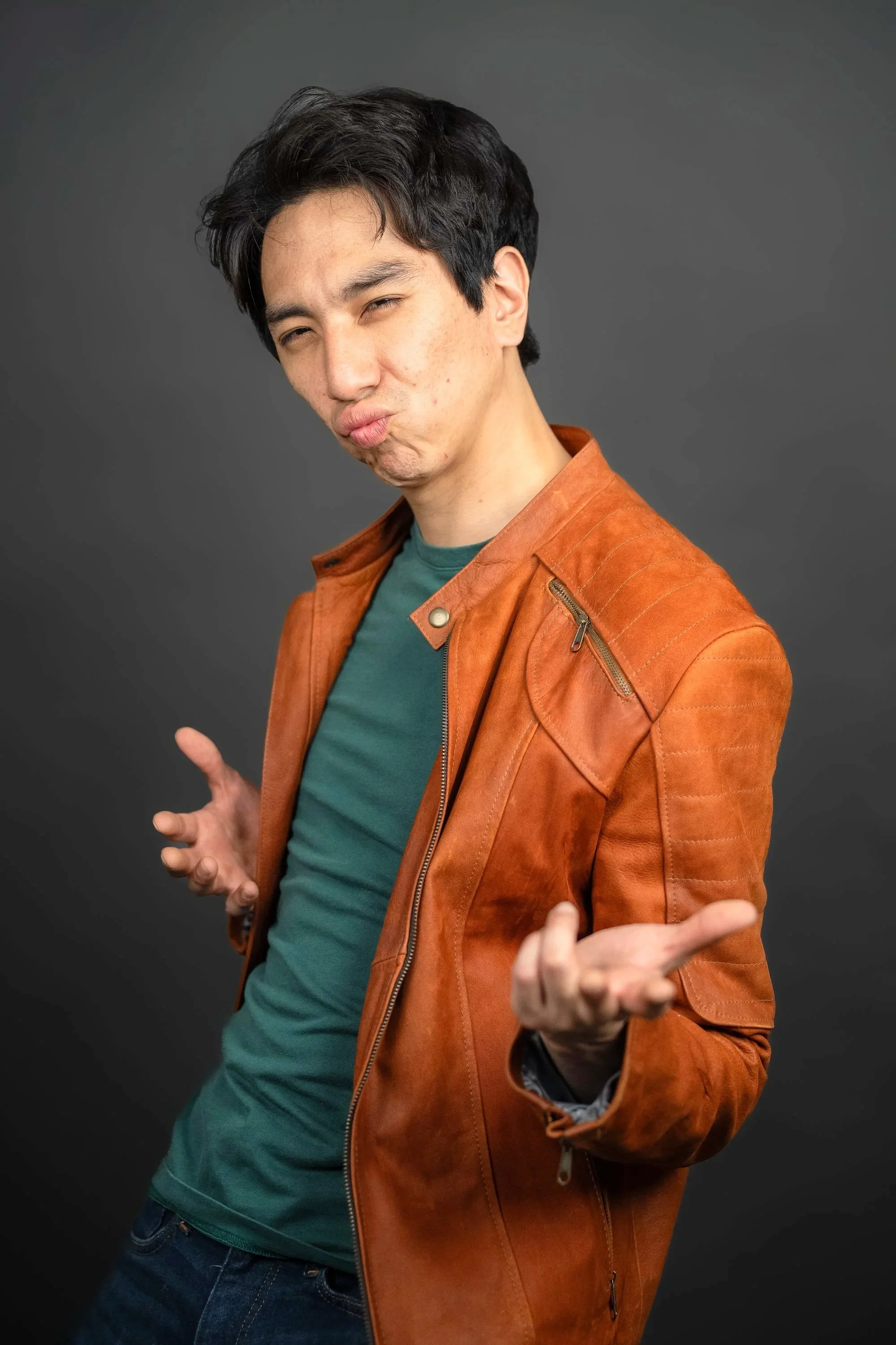 A young man with black hair making a playful, exaggerated pouting expression with his lips. He is wearing a brown leather jacket over a teal shirt, and dark jeans, standing against a plain gray background with arms extended and palms facing upward.