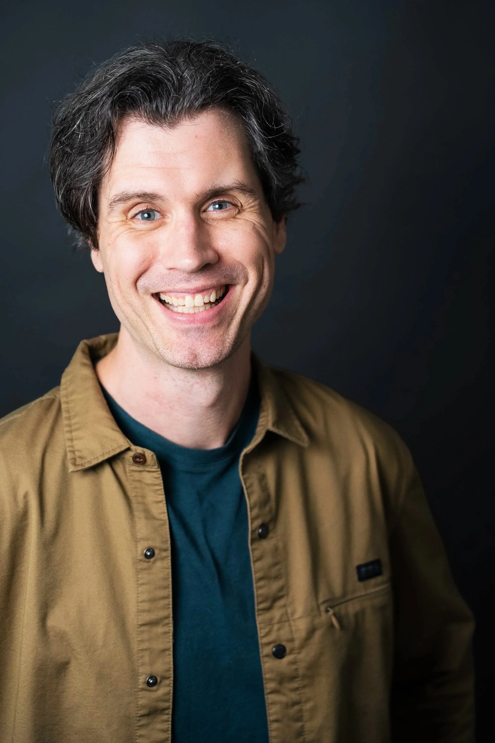 A smiling man with wavy dark hair and blue eyes, wearing a brown jacket over a teal shirt, standing against a dark background.