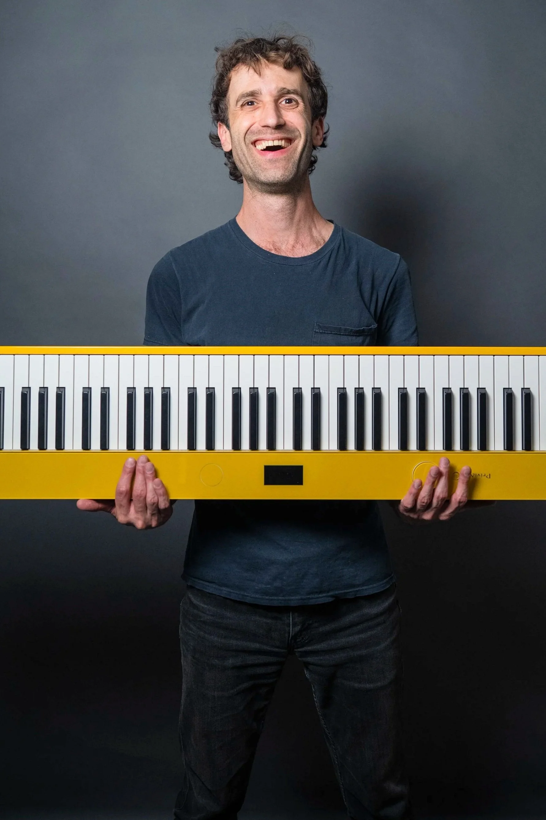 A man with curly dark hair standing against a dark background, smiling and holding a large yellow electronic keyboard in front of him.