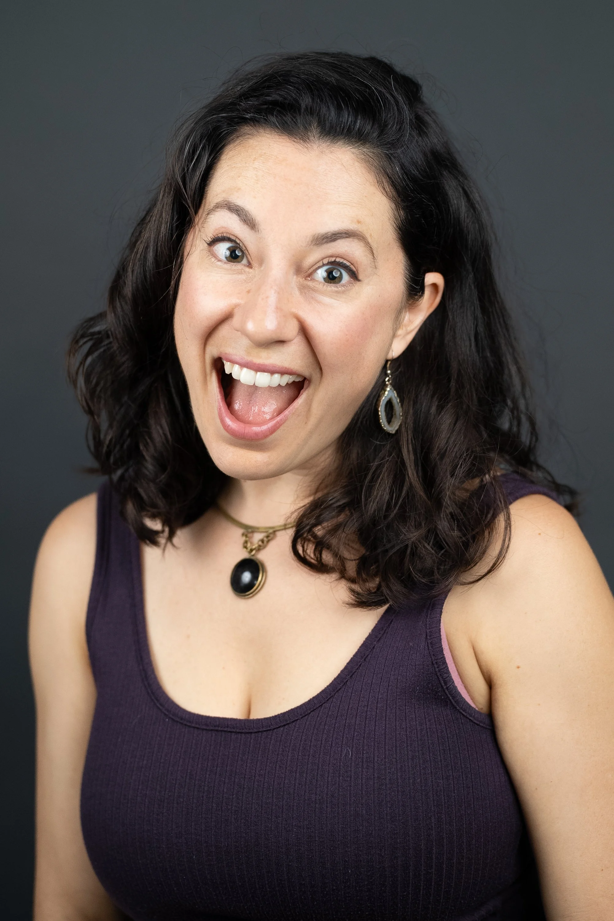 Photo of a woman with dark curly hair, wearing a purple tank top and jewelry, smiling with her mouth open, against a dark gray background.