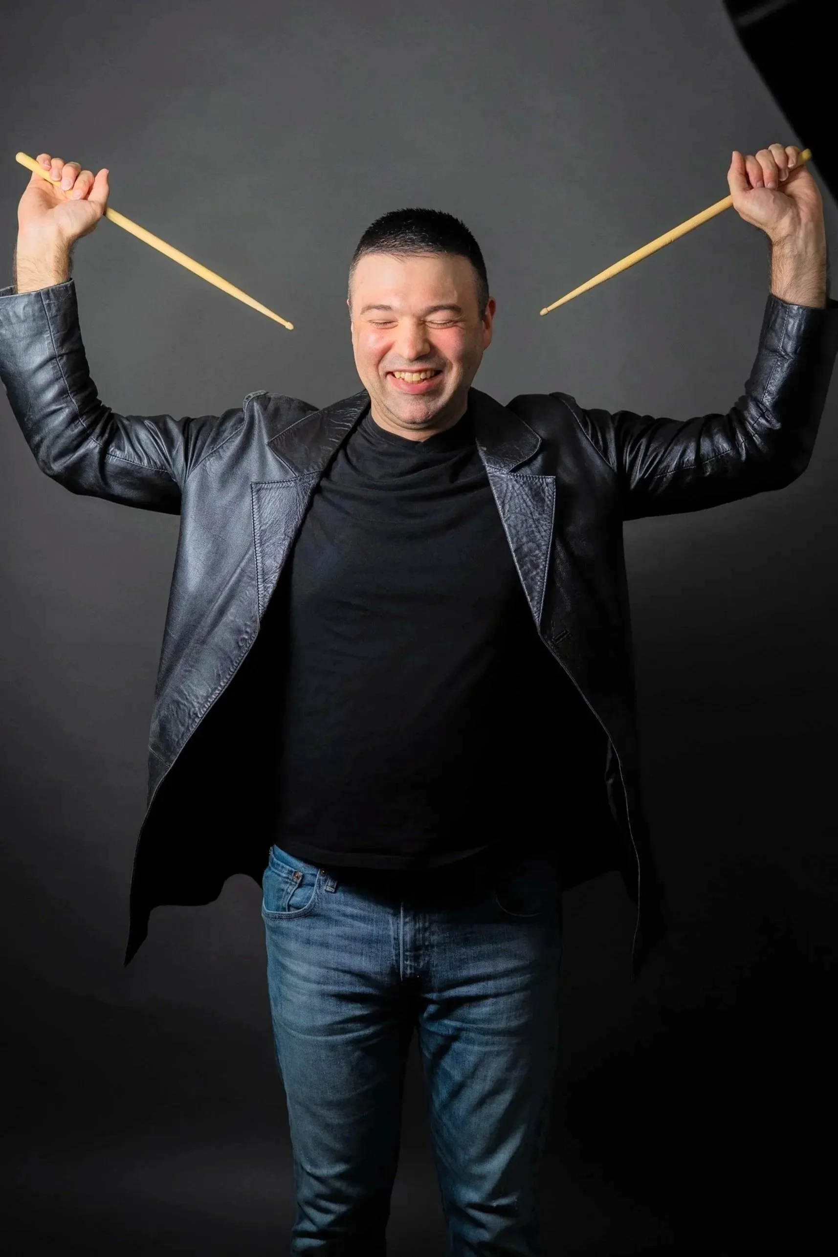 A man with short dark hair, wearing a black leather jacket and blue jeans, is smiling with his eyes closed while holding two drumsticks in the air against a dark gray background.