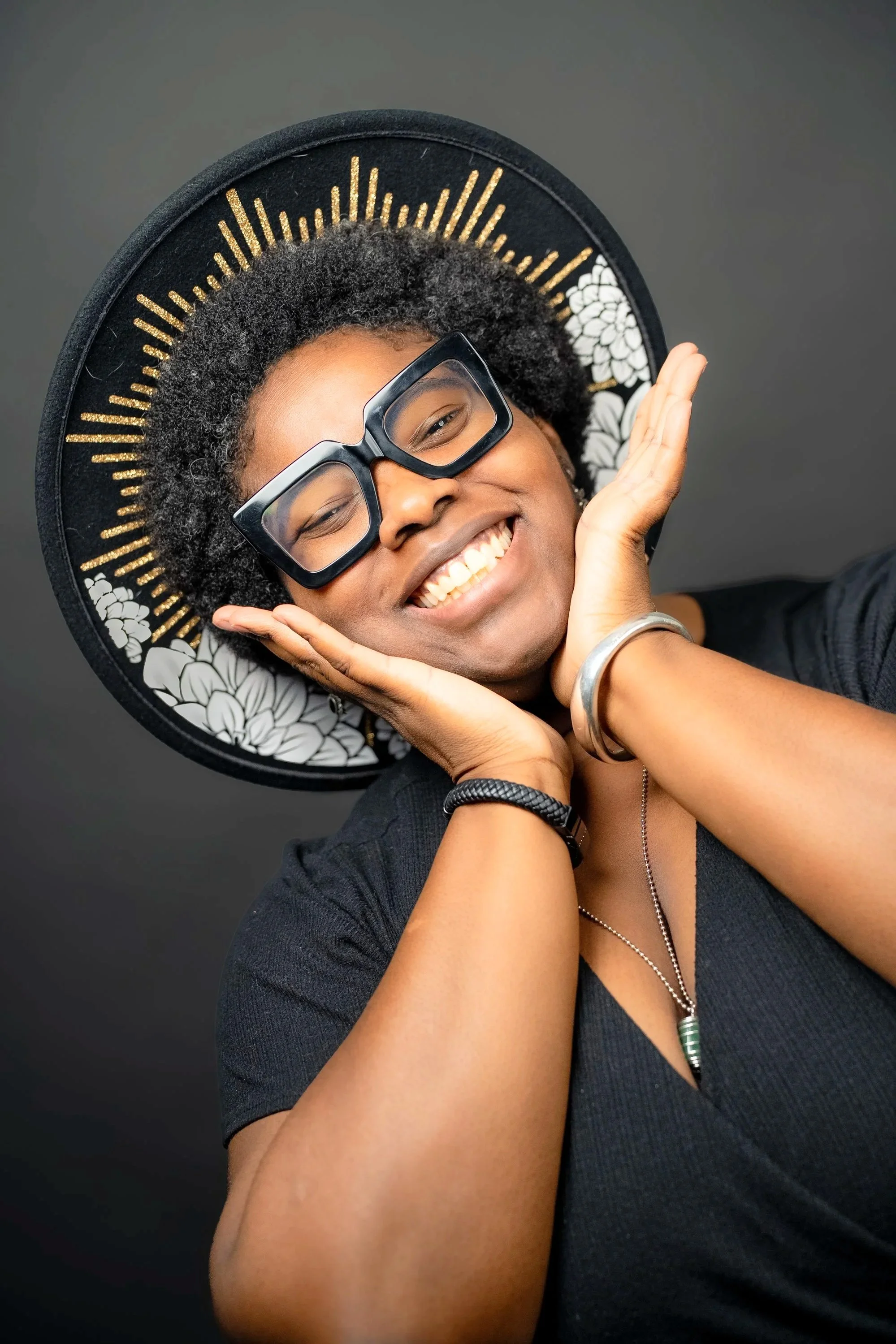 A woman smiling with her hands on her cheeks, wearing large glasses and a wide-brimmed black hat with gold sunburst embellishments.