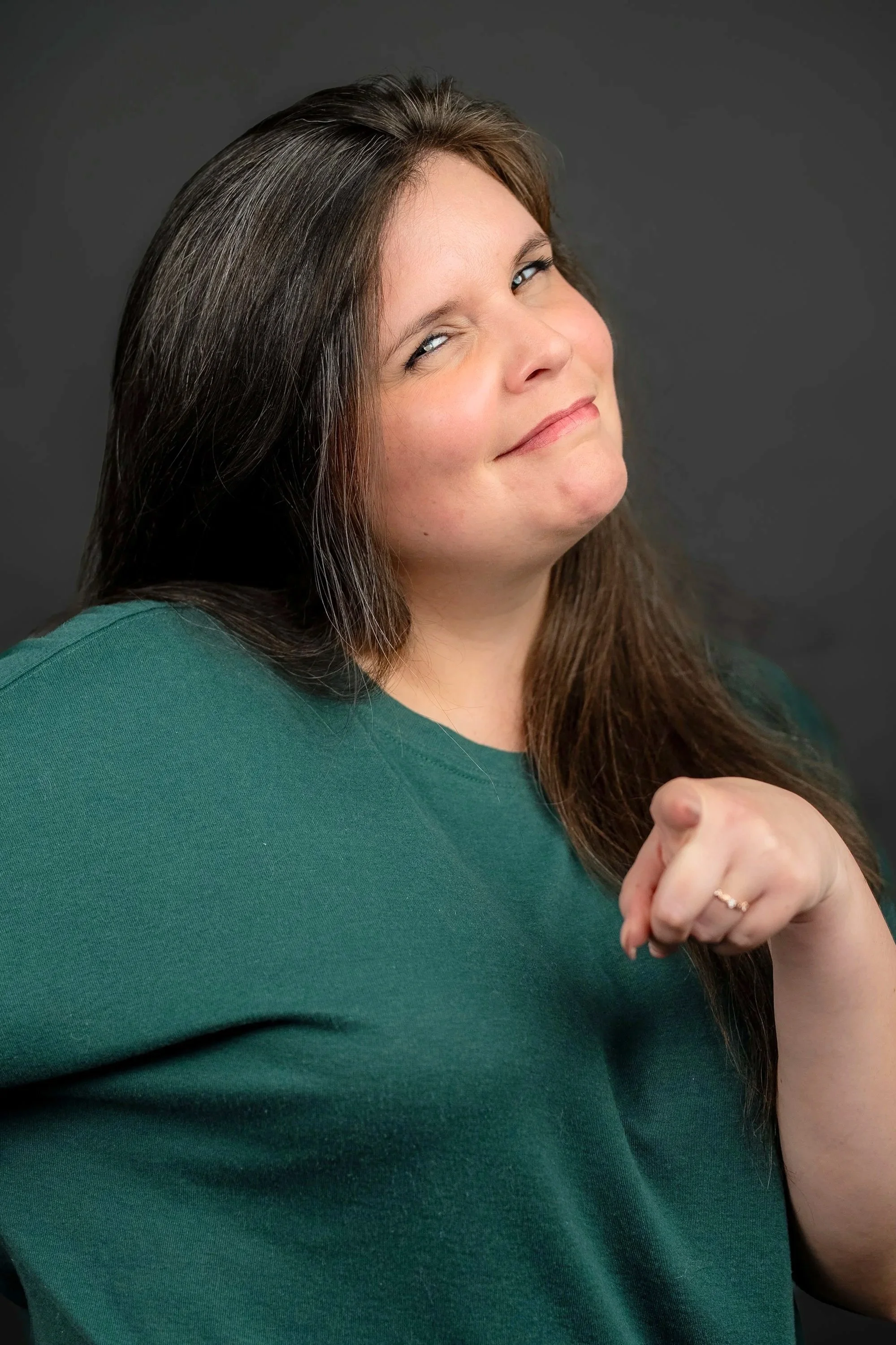 Close-up of a woman with long brown hair, smiling confidently, wearing a dark green top, against a dark background.