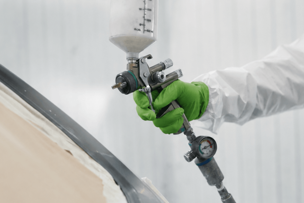 Vehicle painter wearing green gloves using a spray gun to refinish a car in a professional automotive paint booth.