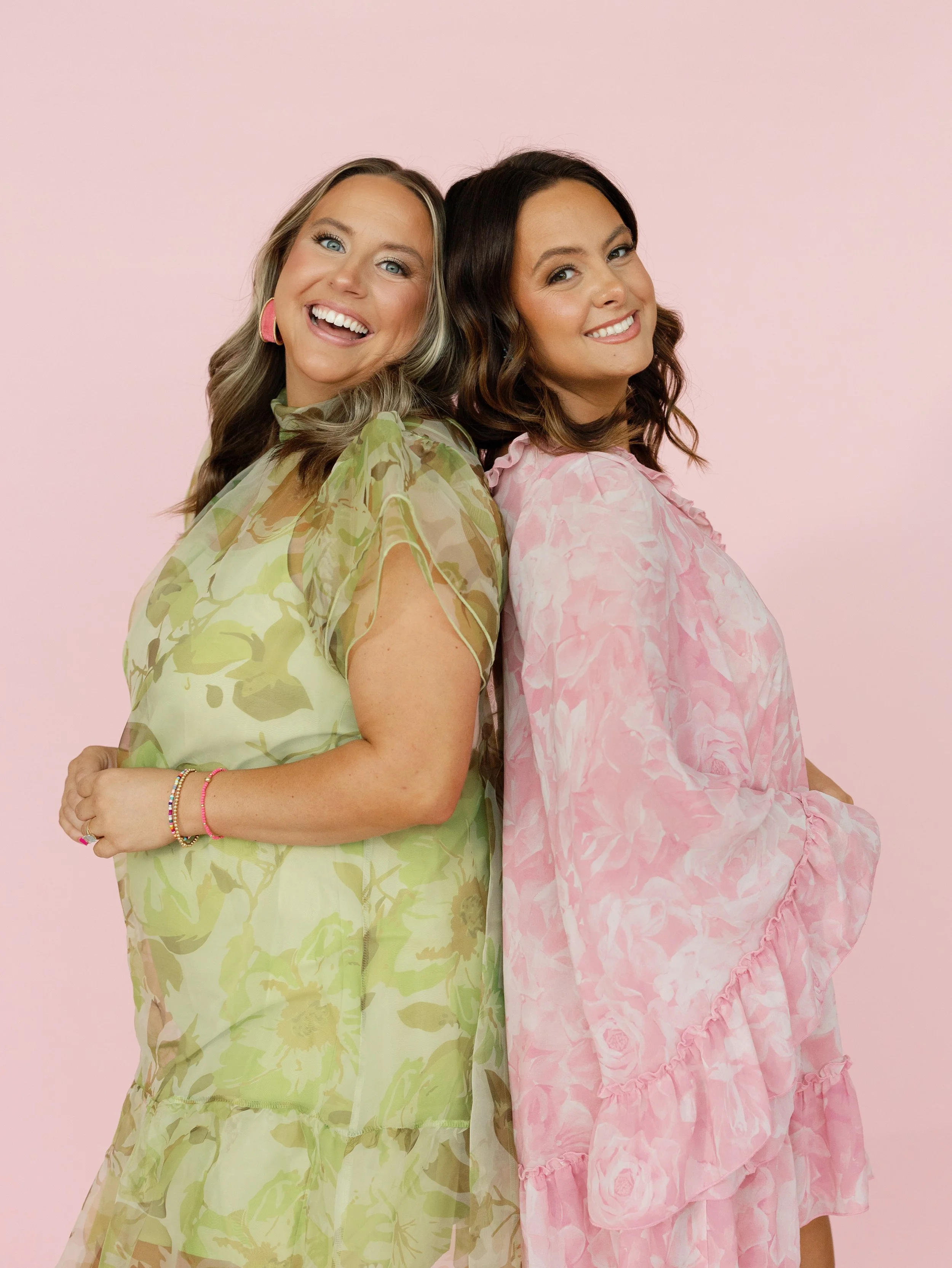 Oklahomas best wedding planners standing back to back, smiling, against a pink background. One is wearing a green floral dress, and the other is in a pink floral dress.