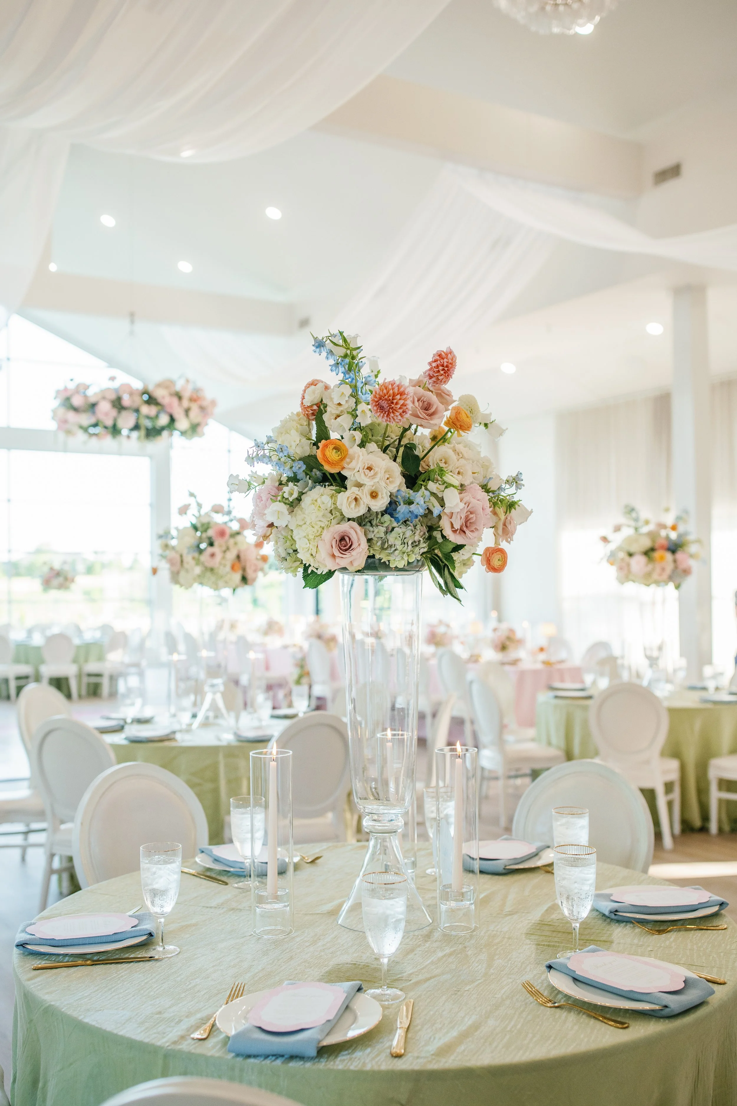 Spring wedding in Tulsa, wedding reception venue decorated with white drapery and small spotlights. There multiple tables with pastel-colored tablecloths, elegant place settings, pink and white floral centerpieces, and tall glass candle holders.
