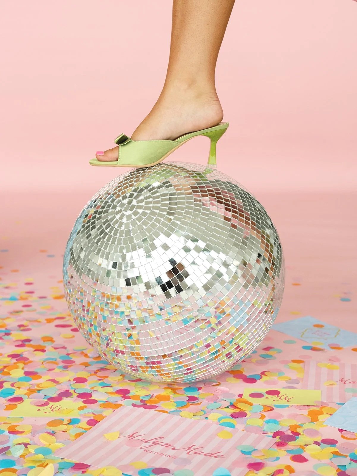 A person's foot wearing a green high-heeled sandal balanced on a large disco ball, with colorful confetti and pink party decorations on the floor, against a pink background.