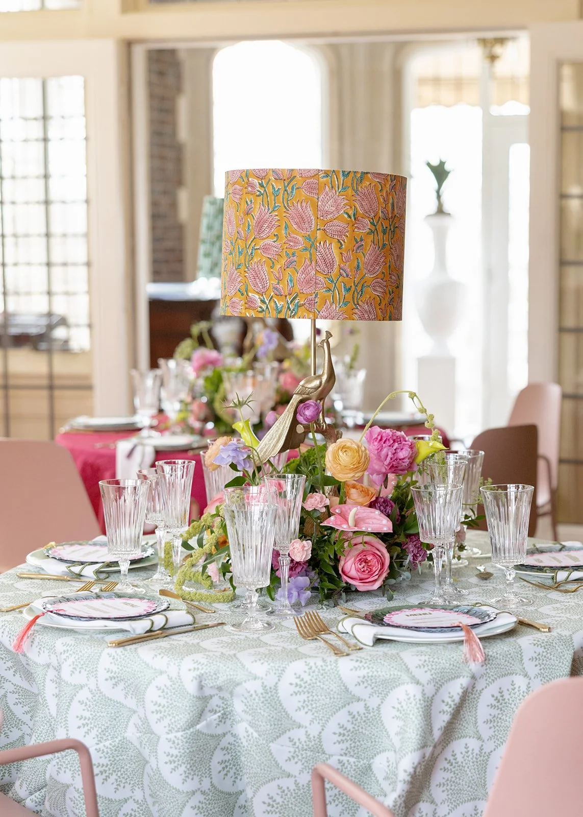 Stunning event in Tulsa Oklahoma, at Harwelden Mansion. Custom details with a  decorated table with pink and floral flowers, glassware, gold cutlery, and napkins, featuring a colorful floral lampshade.