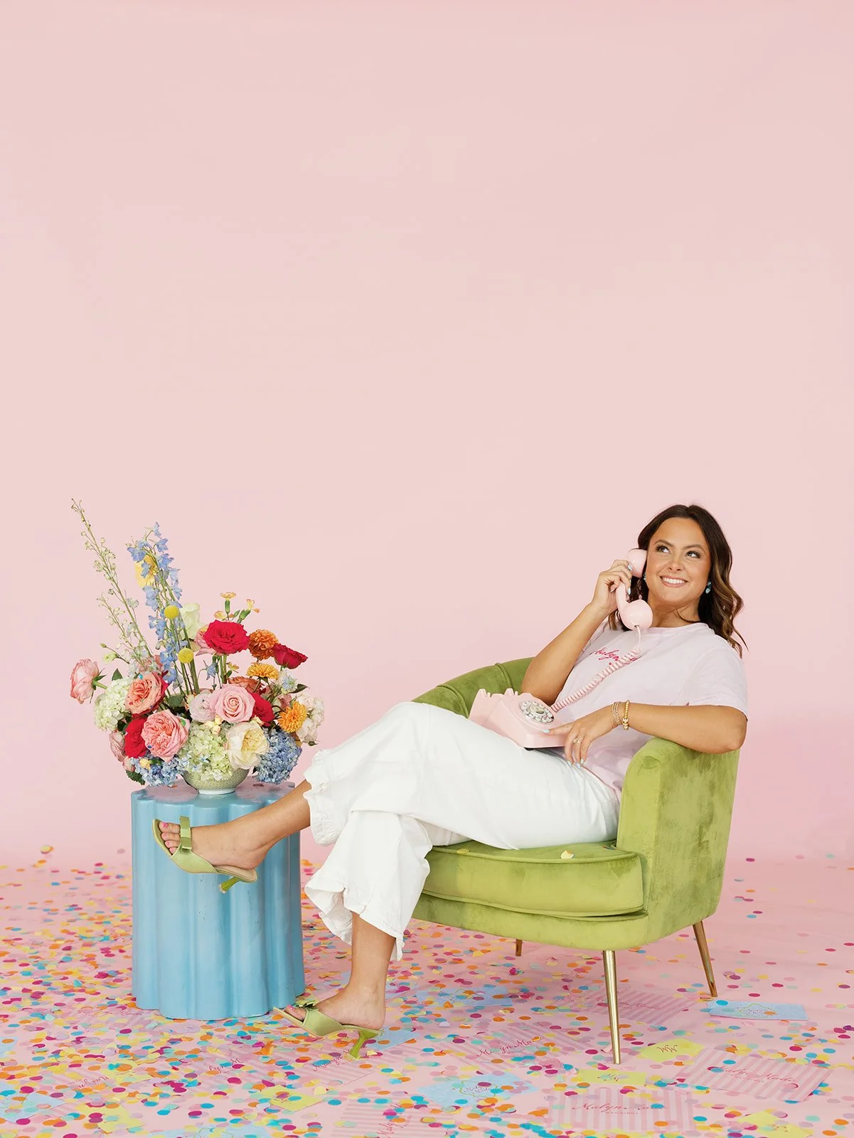 Your luxury wedding planner in Oklahoma! Addie is a woman sitting in a green chair, smiling, holding a pink rotary phone, surrounded by colorful confetti and flowers, with a pink background. Says call us for your event needs