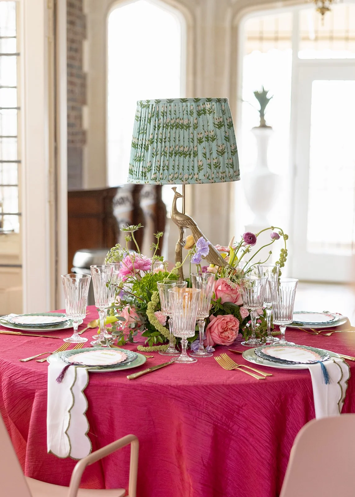 A round dining table with a red tablecloth, set with glassware, plates, and gold utensils. A floral centerpiece with pink and purple flowers holds a vintage-style lampshade with a peacock figurine. The background shows a bright room with large windows and decorative interior elements.